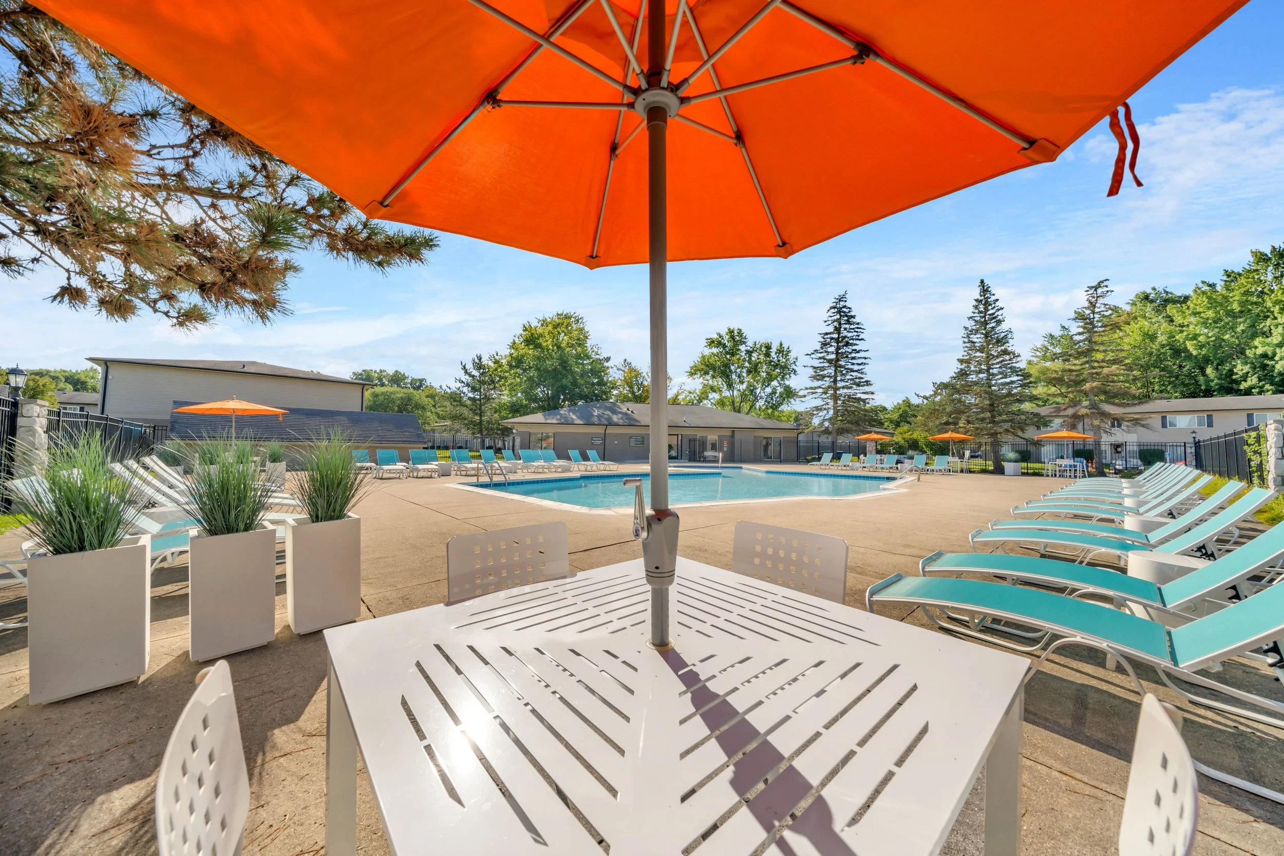 View of a swimming pool area with white lounge chairs, orange umbrellas, and a small white table in the foreground. There are tall trees, a fence, and buildings in the background under a partly cloudy sky.
