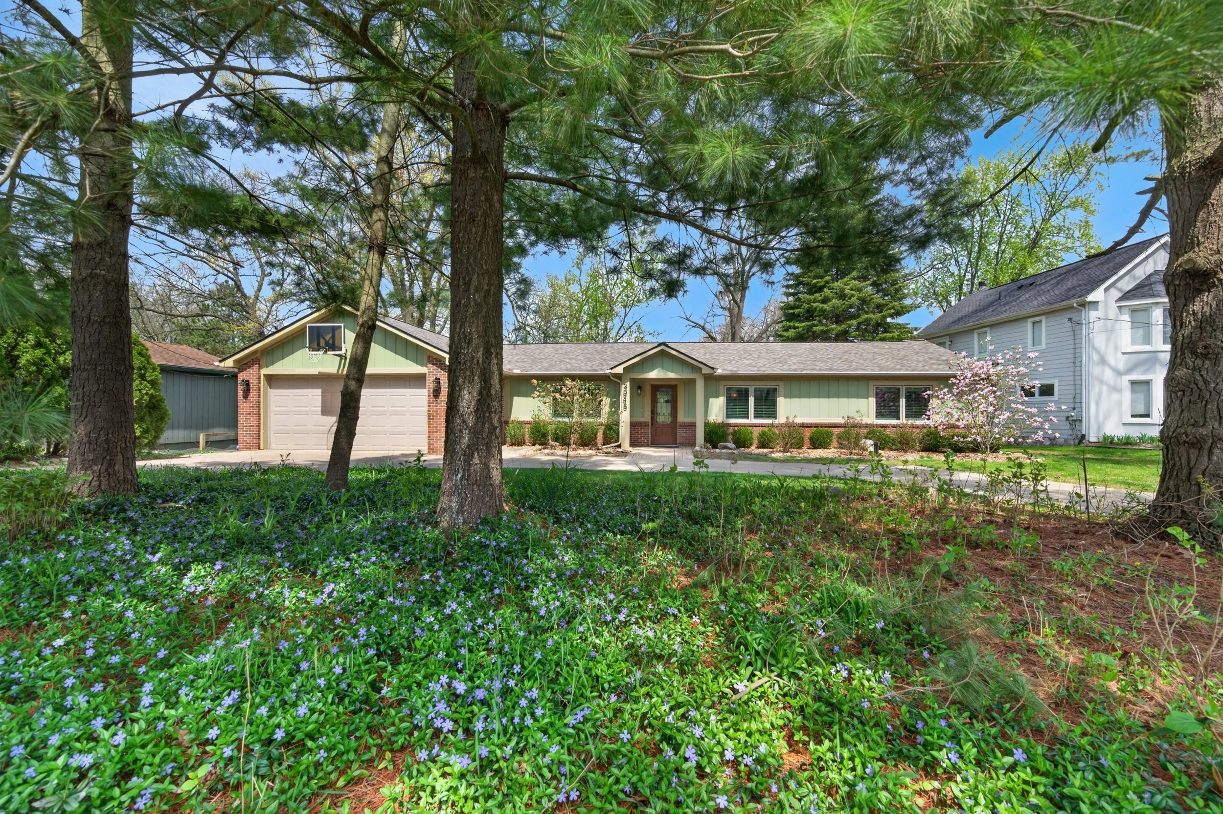 A house with a beige garage door and green siding, surrounded by trees and flowering plants, with a front yard filled with purple flowers and the sky visible through the tree branches.