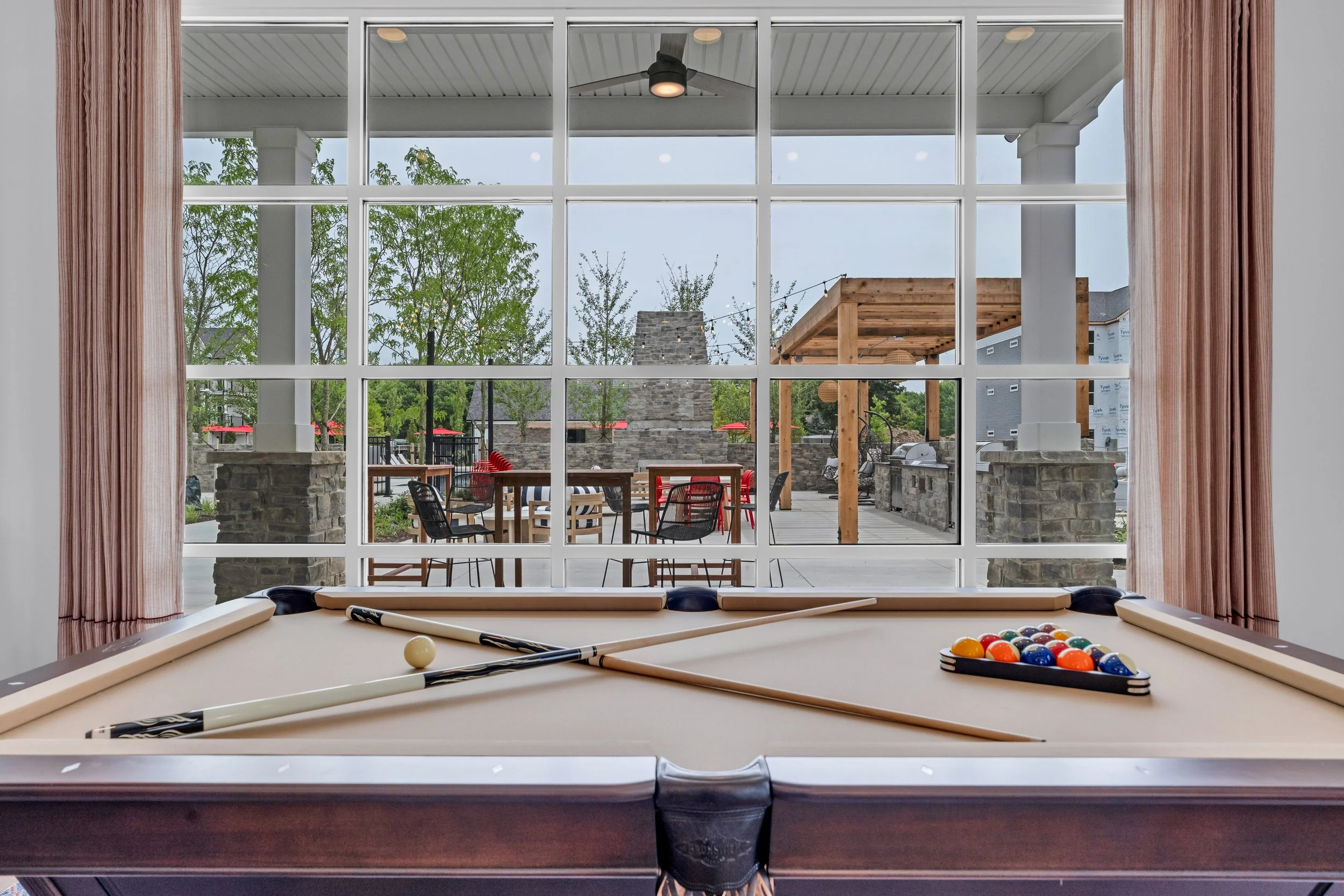 Interior of a room with a pool table and a large window view of an outdoor patio with a barbecue area, trees, and outdoor furniture.