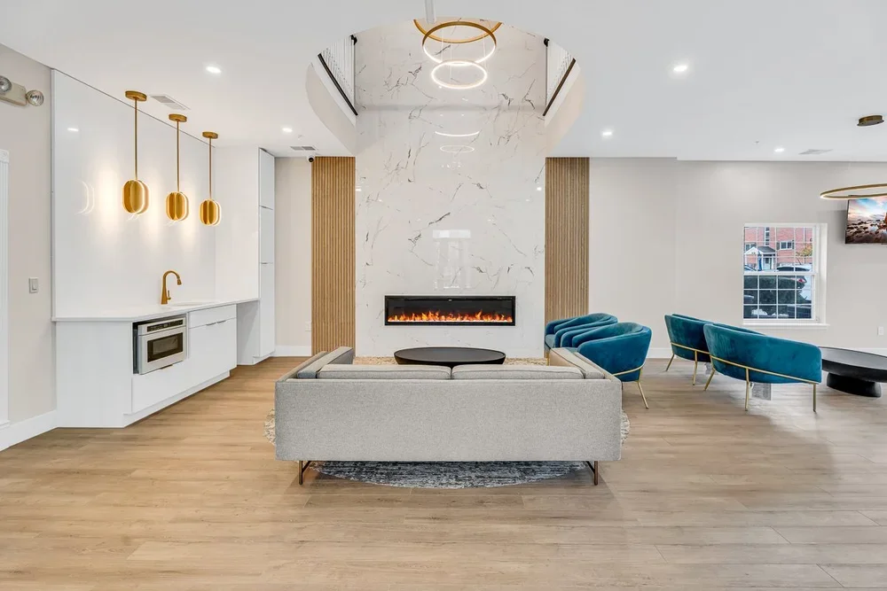 Modern living room with a white marble accent wall, fireplace, gray sofa, blue velvet chairs, wooden flooring, and a kitchenette with gold pendant lights.