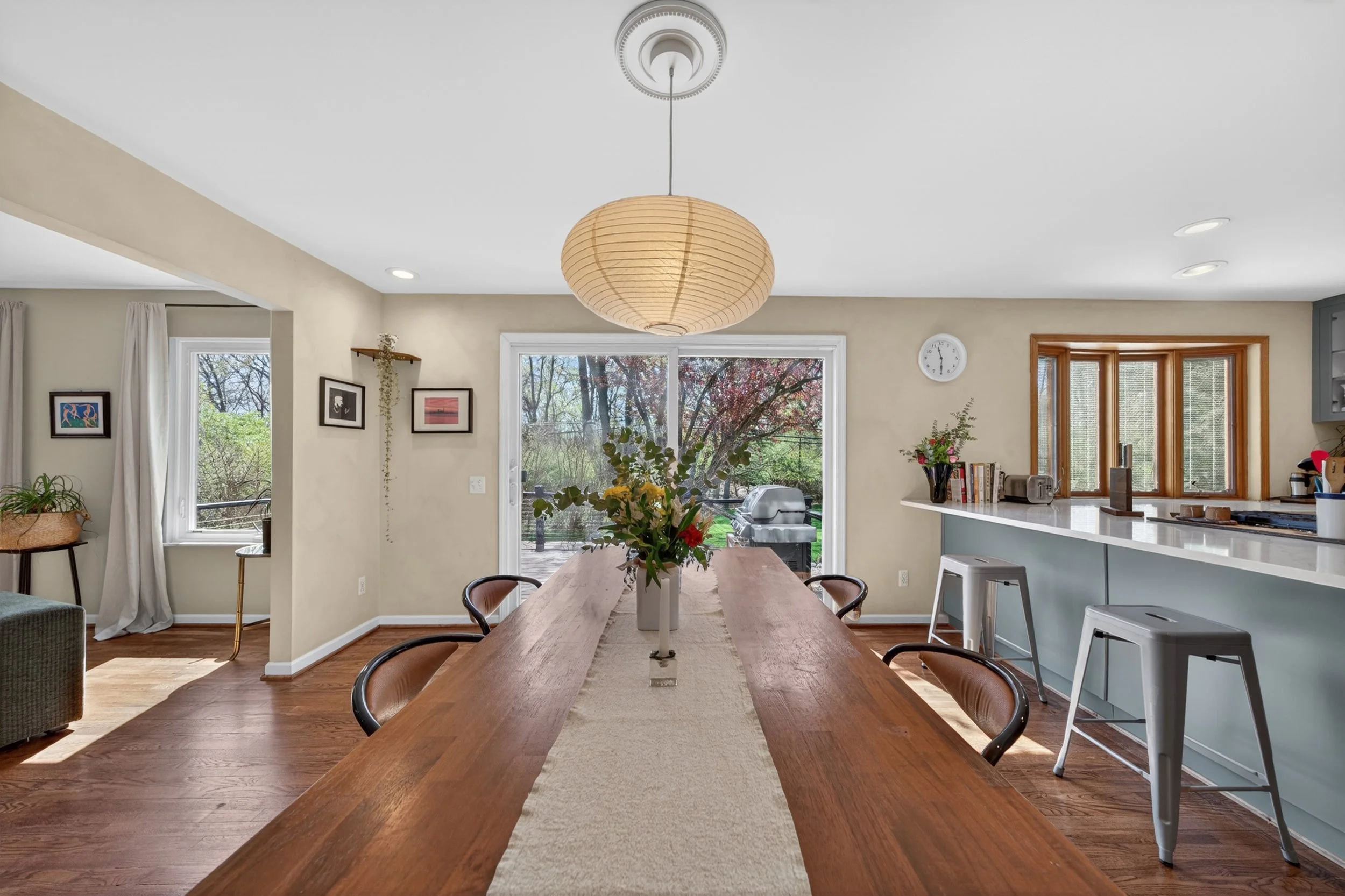 A bright dining area with a long wooden table, black chairs, a vase of flowers, large windows showing trees outside, framed pictures on the wall, and a view of the kitchen with a window and countertop.