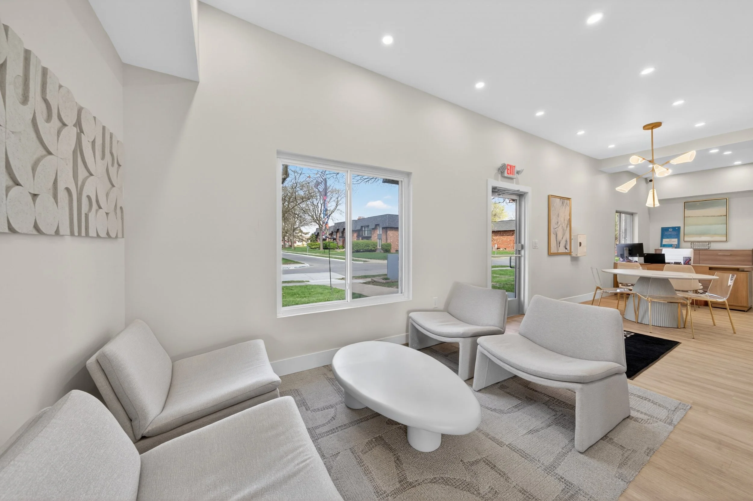 A lobby area with white walls, large windows, modern white furniture, a wooden desk, and a ceiling fan with multiple lights.