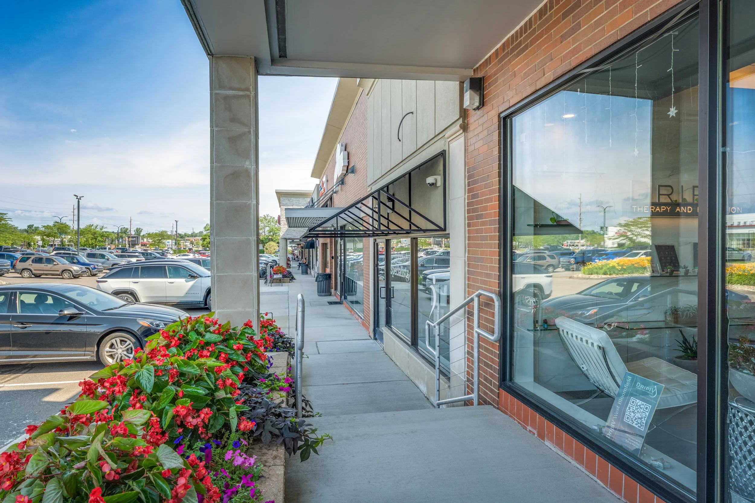 Sidewalk view of a shopping plaza with parked cars, colorful flowers, and storefronts with large glass windows, some with signs and doorways.