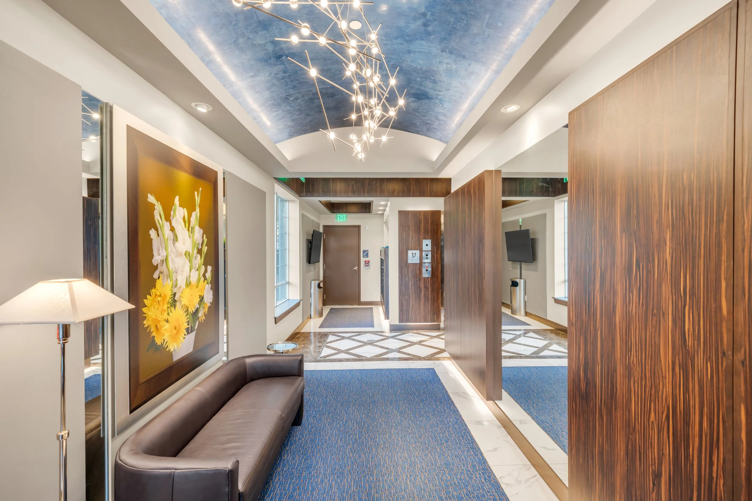 Interior of a modern lobby with a blue painted ceiling, a contemporary chandelier, wooden accents, a floral painting, a sofa, and an exit door.