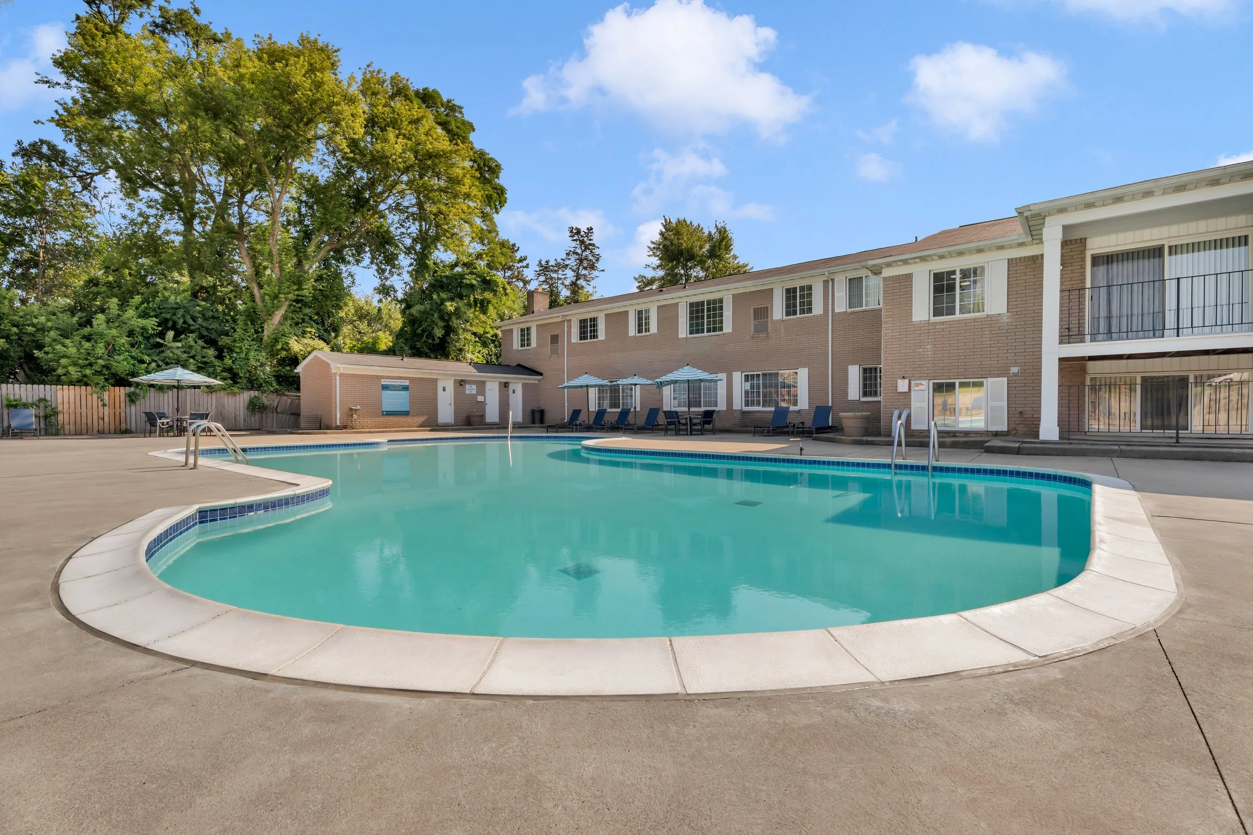 An outdoor swimming pool area with lounge chairs and umbrellas, surrounded by a beige brick apartment building and green trees under a partly cloudy sky.