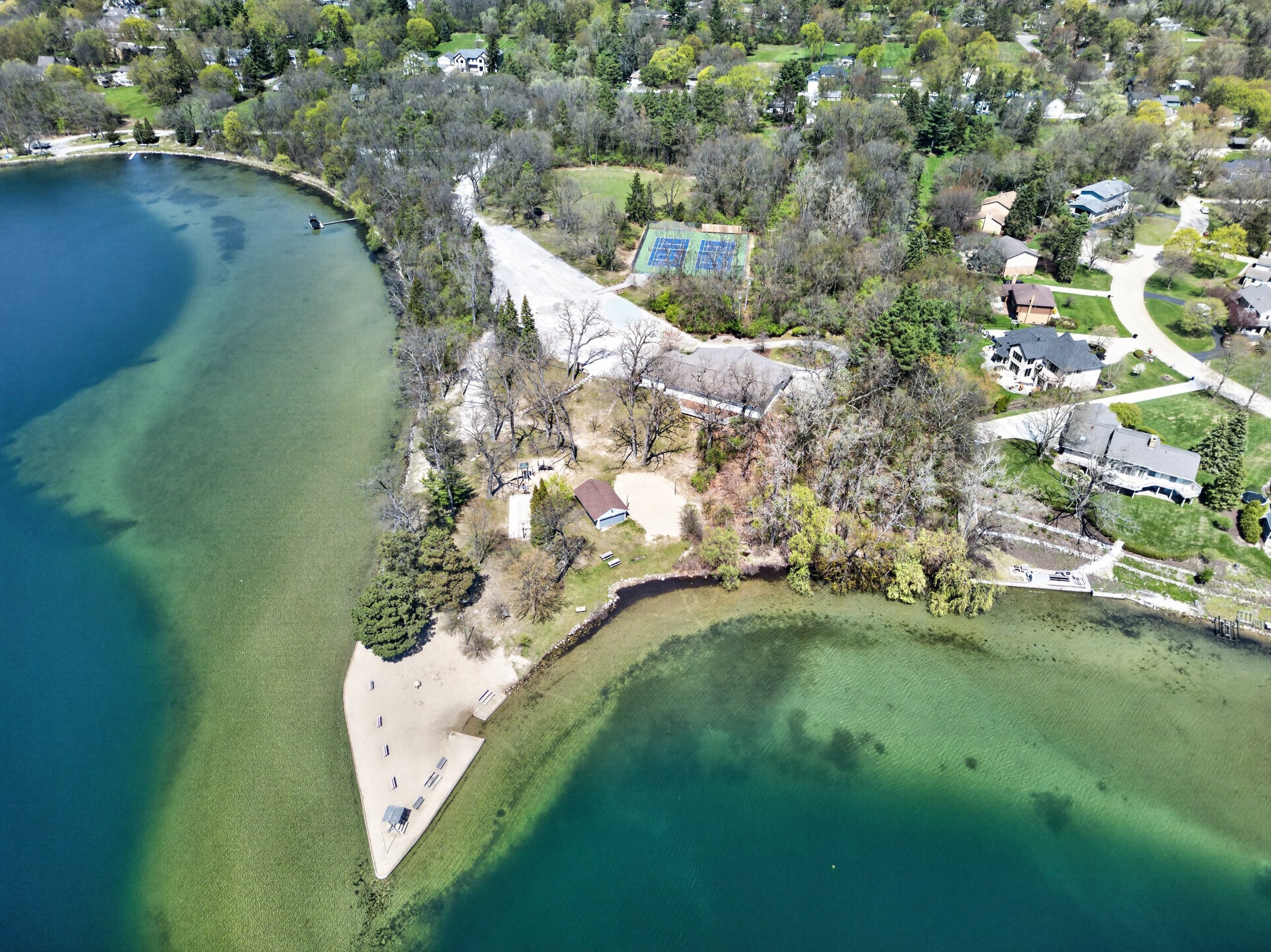 Aerial view of a lakeside park with a sandy beach, picnic tables, and walking paths, surrounded by residential houses and trees.