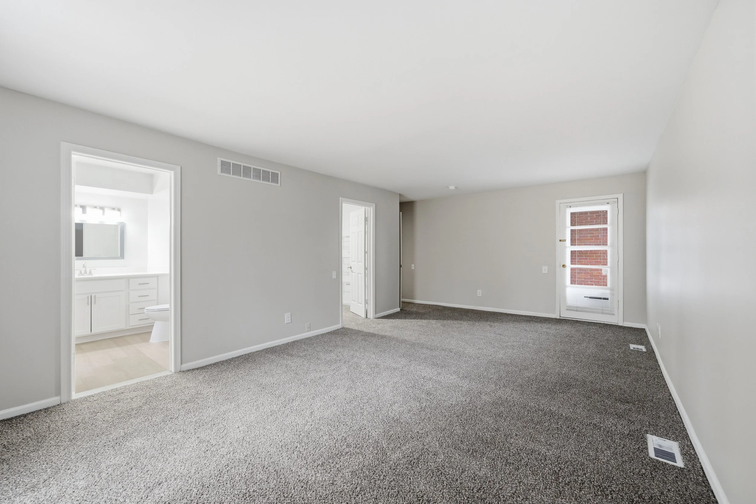 Unfurnished living room with light gray walls, carpeted floor, and white trim. Features a large window, a door leading outside, and an open doorway to a bathroom.