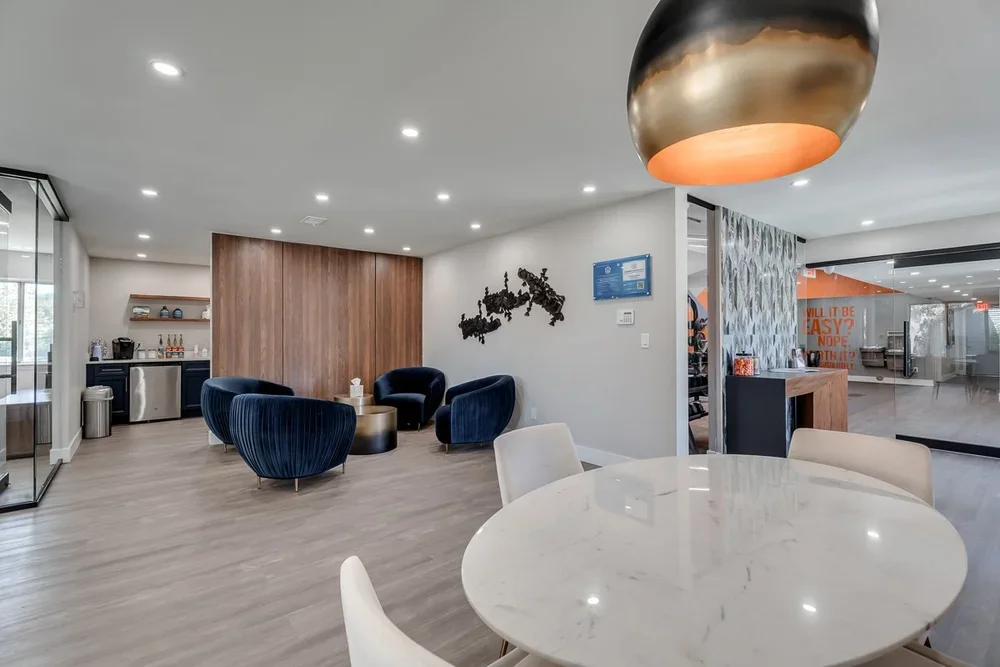 Modern hotel lobby with blue armchairs, round coffee table, and a kitchenette area with coffee maker, refrigerator, and snacks. Recessed ceiling lights and a contemporary metallic pendant light. Artwork on the wall and a wooden panel background.