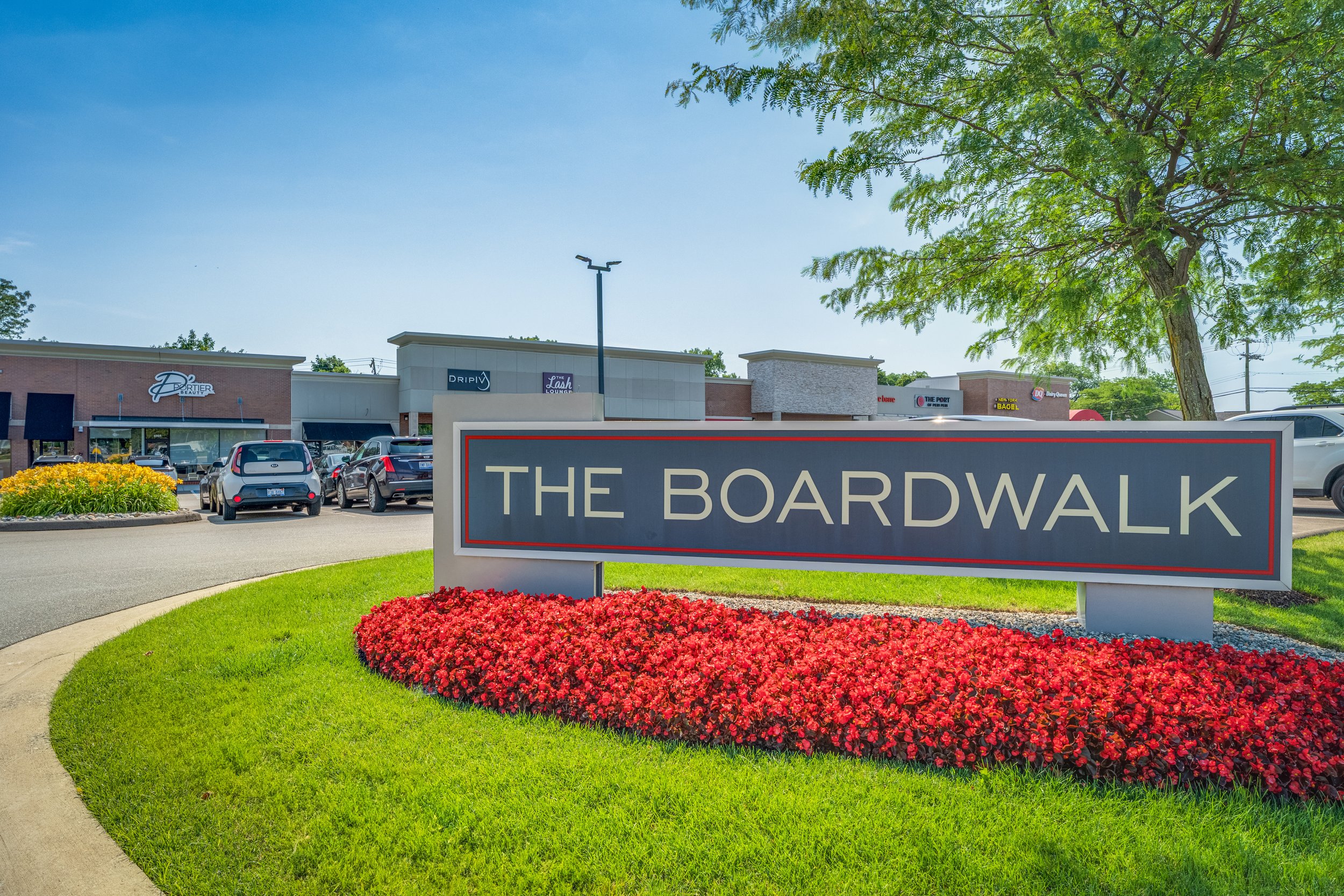 Sign reading 'The Boardwalk' in front of a shopping center with parked cars and stores, surrounded by neatly trimmed grass and red flowers, under a blue sky with green trees.