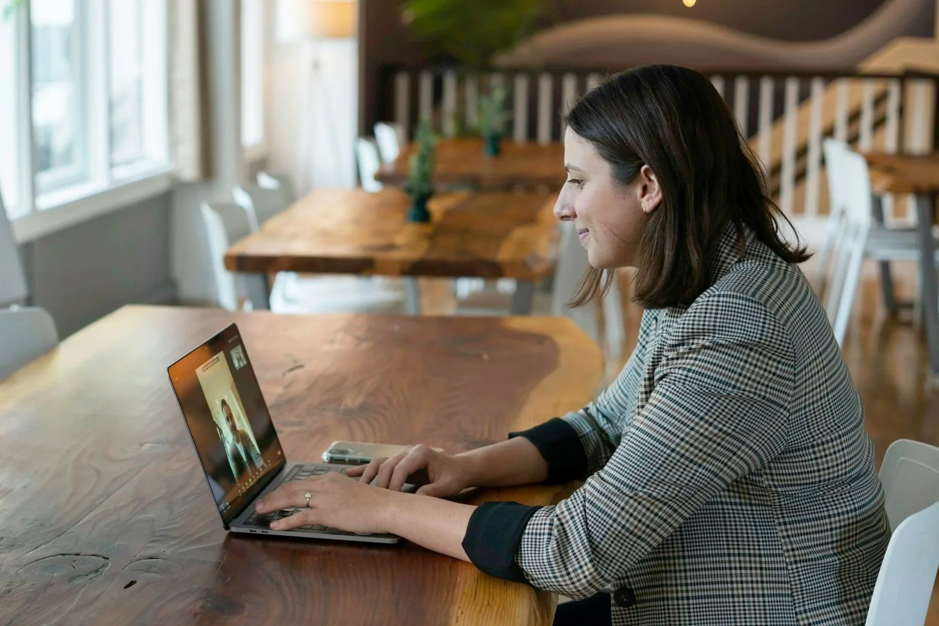 A woman sitting at a wooden table using a laptop while on a video call in a well-lit room with large windows.