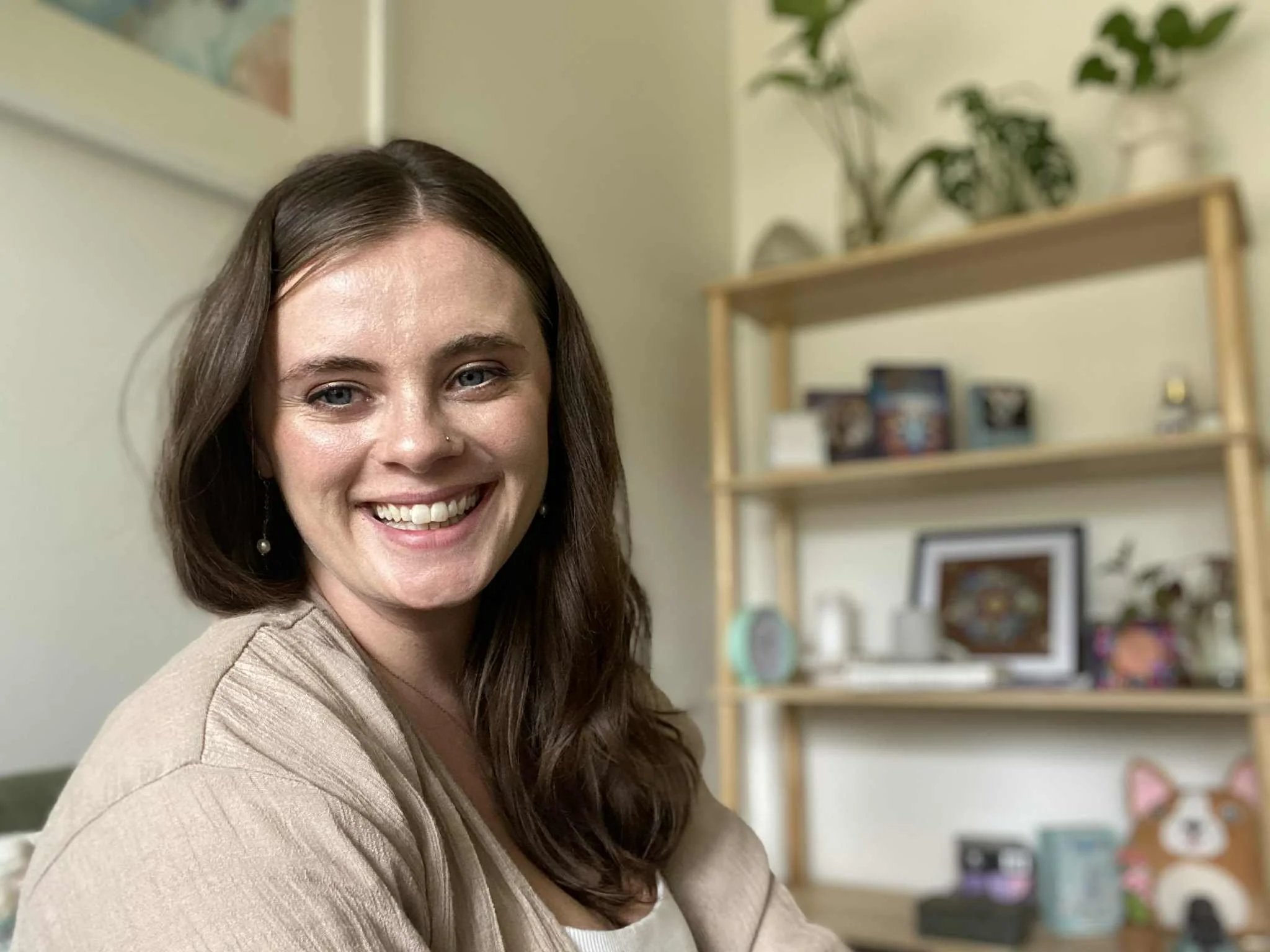 Smiling woman with brown hair and earrings in a room with a wooden shelf and various decorative items.