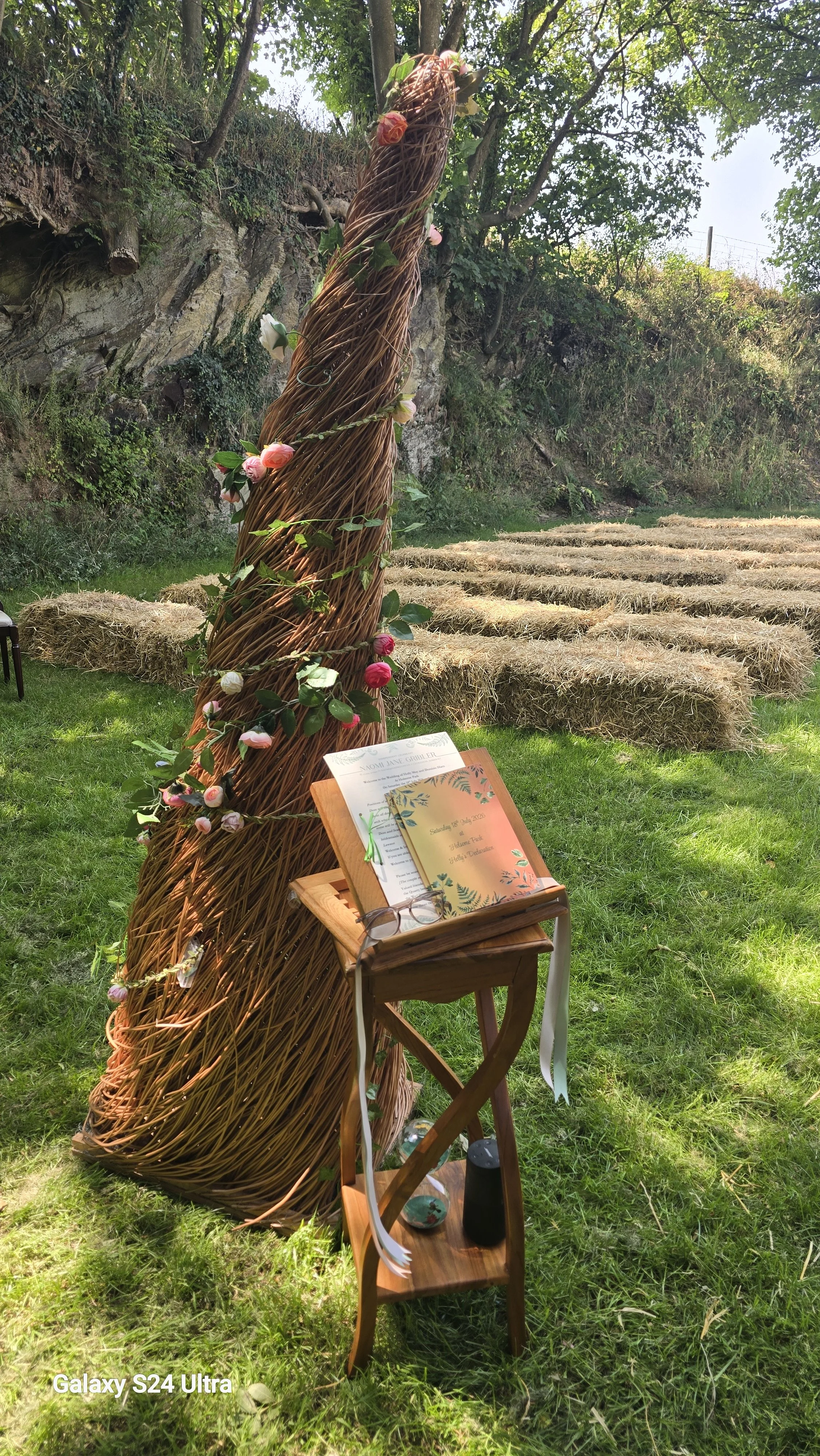 Decorative outdoor wedding setup featuring a triangular wicker arch adorned with pink and white roses and green leaves, a small wooden table with an open guest book and reading glasses, and hay bales in a grassy area surrounded by trees.