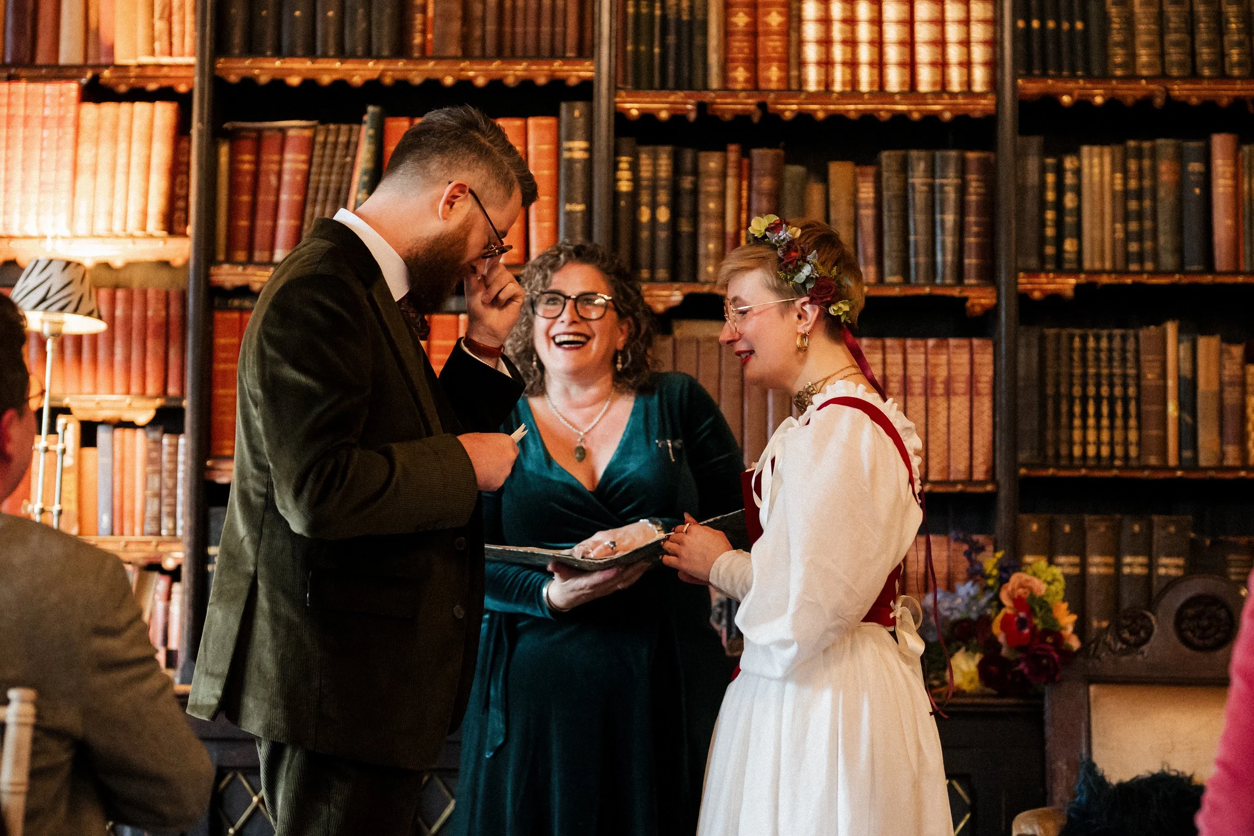 A wedding ceremony taking place in the gorgeous library  at Huntsham Court in Devon. The Bride and Groom and NaomiJane the celebrant are really enjoying this intimate moment when they exchange vows.