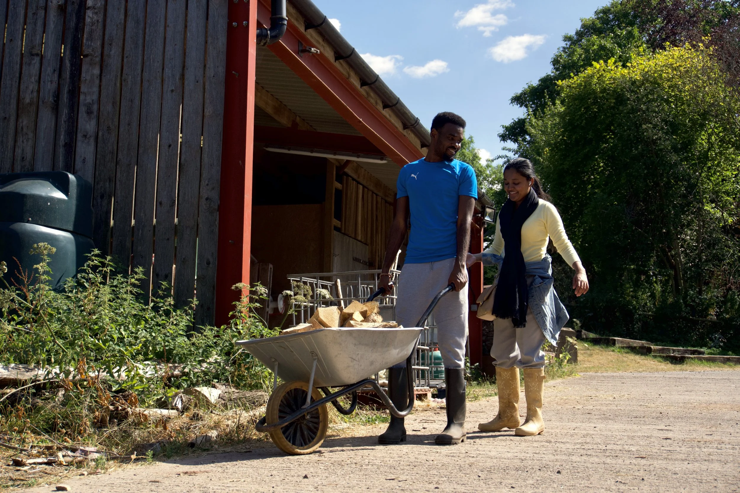 Two people are working in a garden beside a building with dark wooden siding and a red metal roof. They are both wearing rubber boots and casual clothing, and they are smiling. One person is pushing a wheelbarrow filled with chunks of wood, while the other appears to be holding a gardening tool. There are lush green trees and plants in the background, and the sky is blue with a few white clouds.