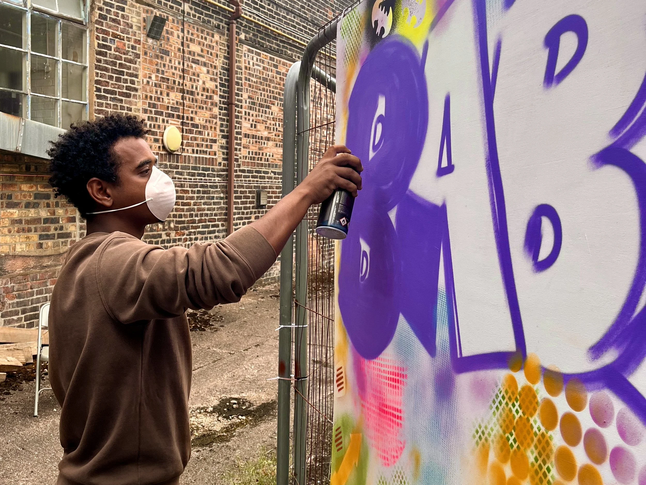 A man wearing a white face mask and a brown hoodie spray paints a colorful mural on a large white wall. The mural includes vibrant colors and abstract patterns. The setting appears to be outdoors in an urban area with brick buildings and metal fencing in the background.