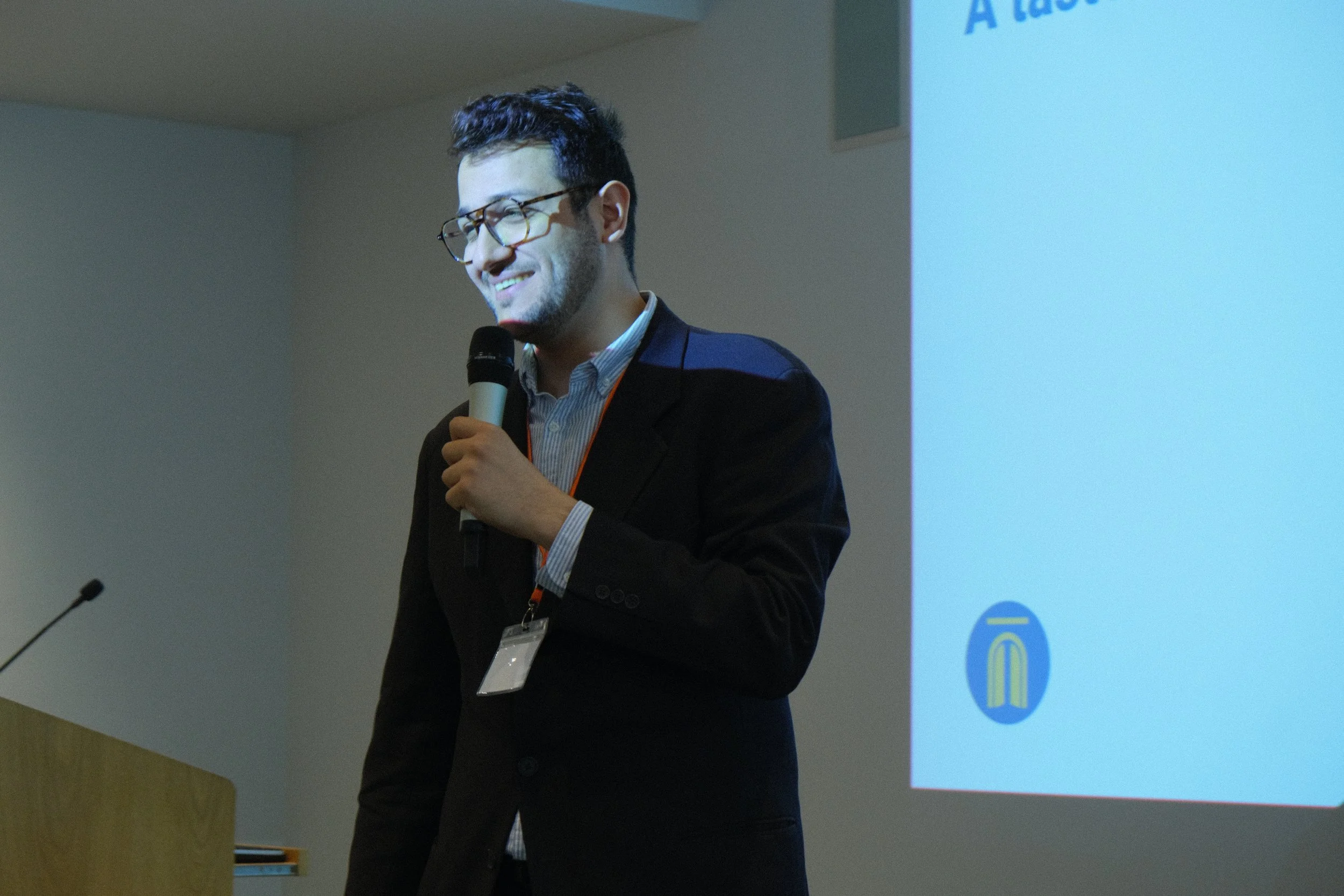 A man with dark hair and glasses, wearing a dark suit, standing in front of a presentation screen and smiling while holding a microphone.