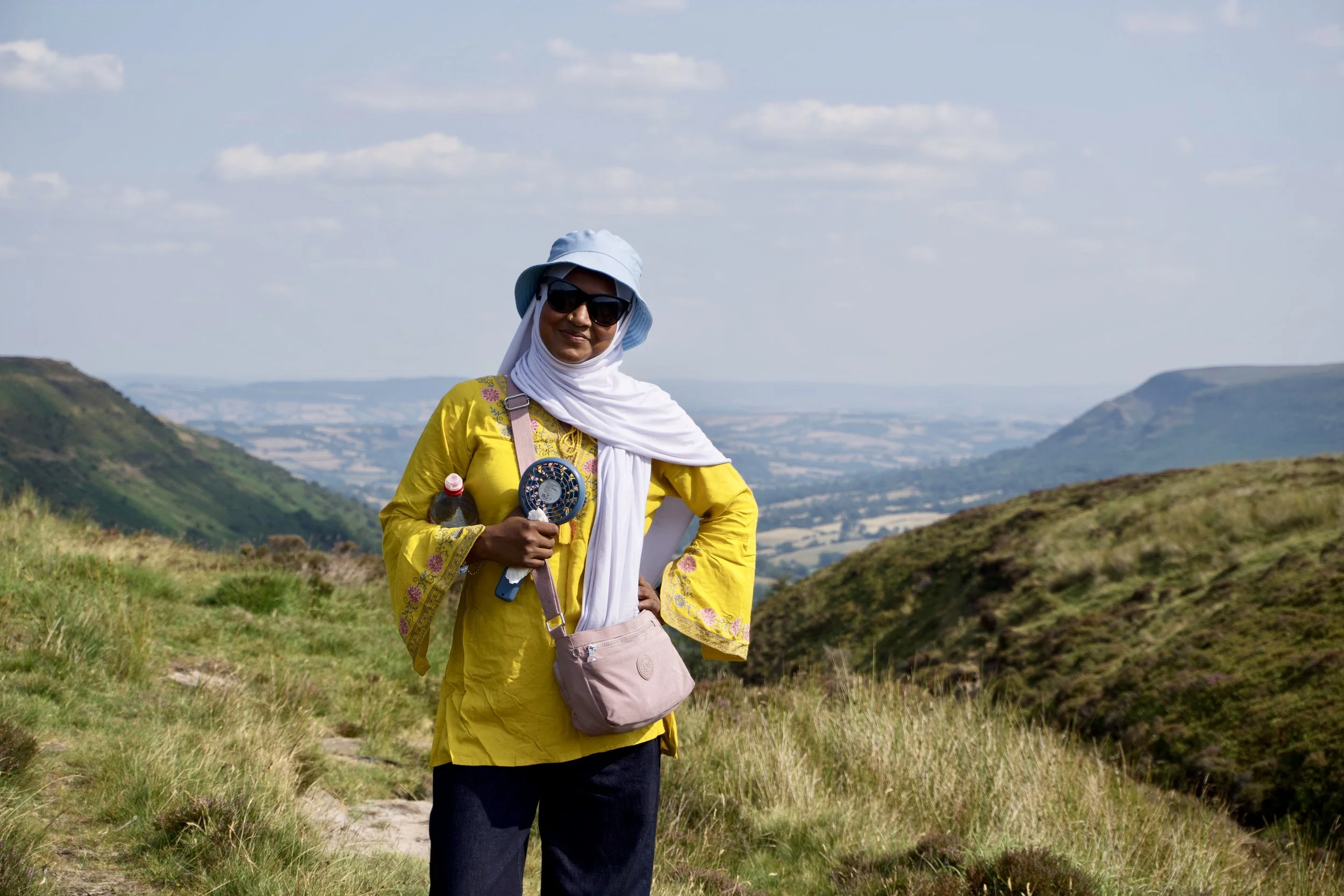 Woman in a yellow jacket, white scarf, black sunglasses, and hat standing outdoors in a scenic hilly landscape, holding a water bottle and other small items.
