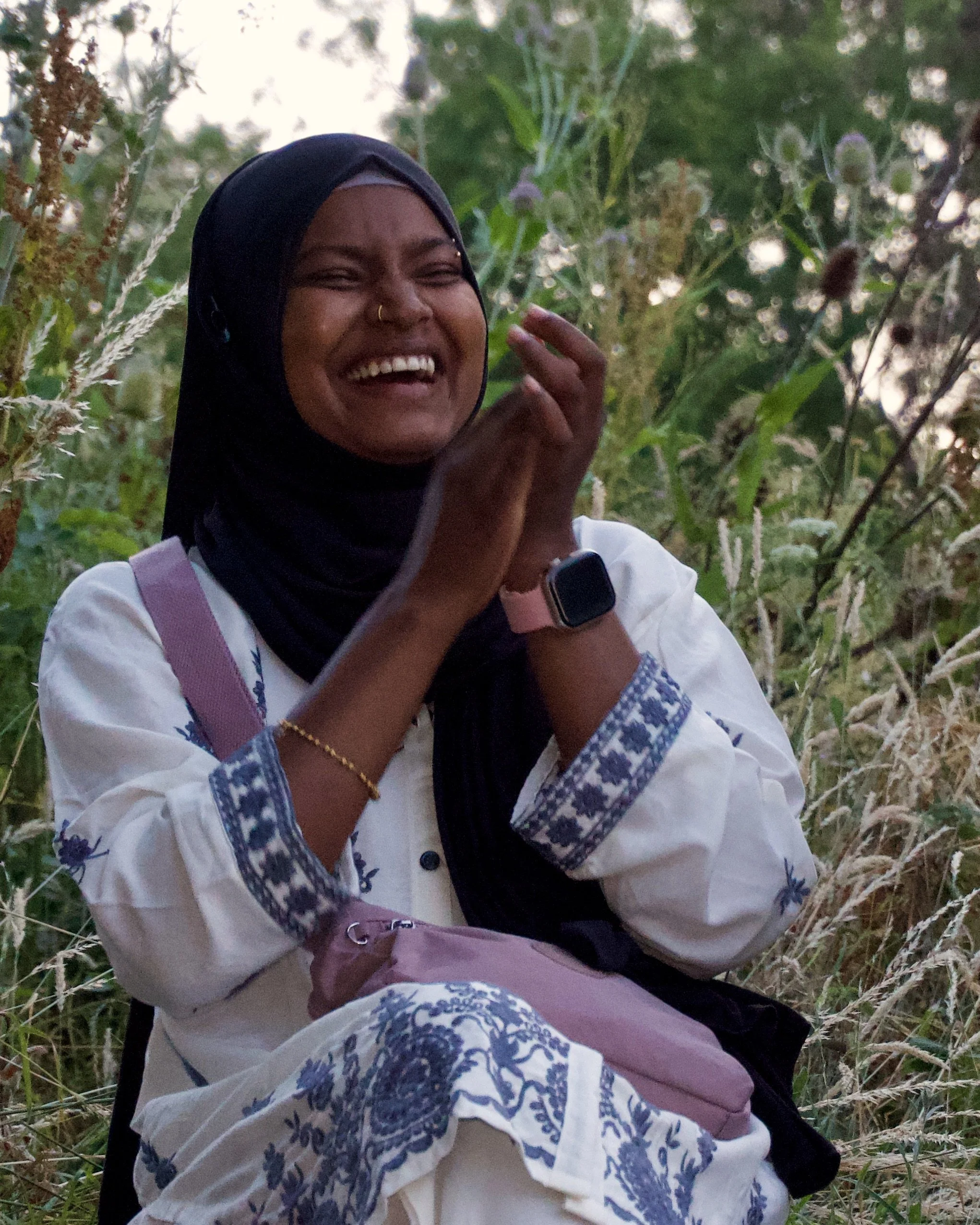 A woman with dark skin and wearing a black hijab, smiling and laughing, sitting outdoors among tall green grass and plants, wearing a white shirt with blue embroidery and a pink strap, possibly a bag strap, and a smartwatch on her wrist.