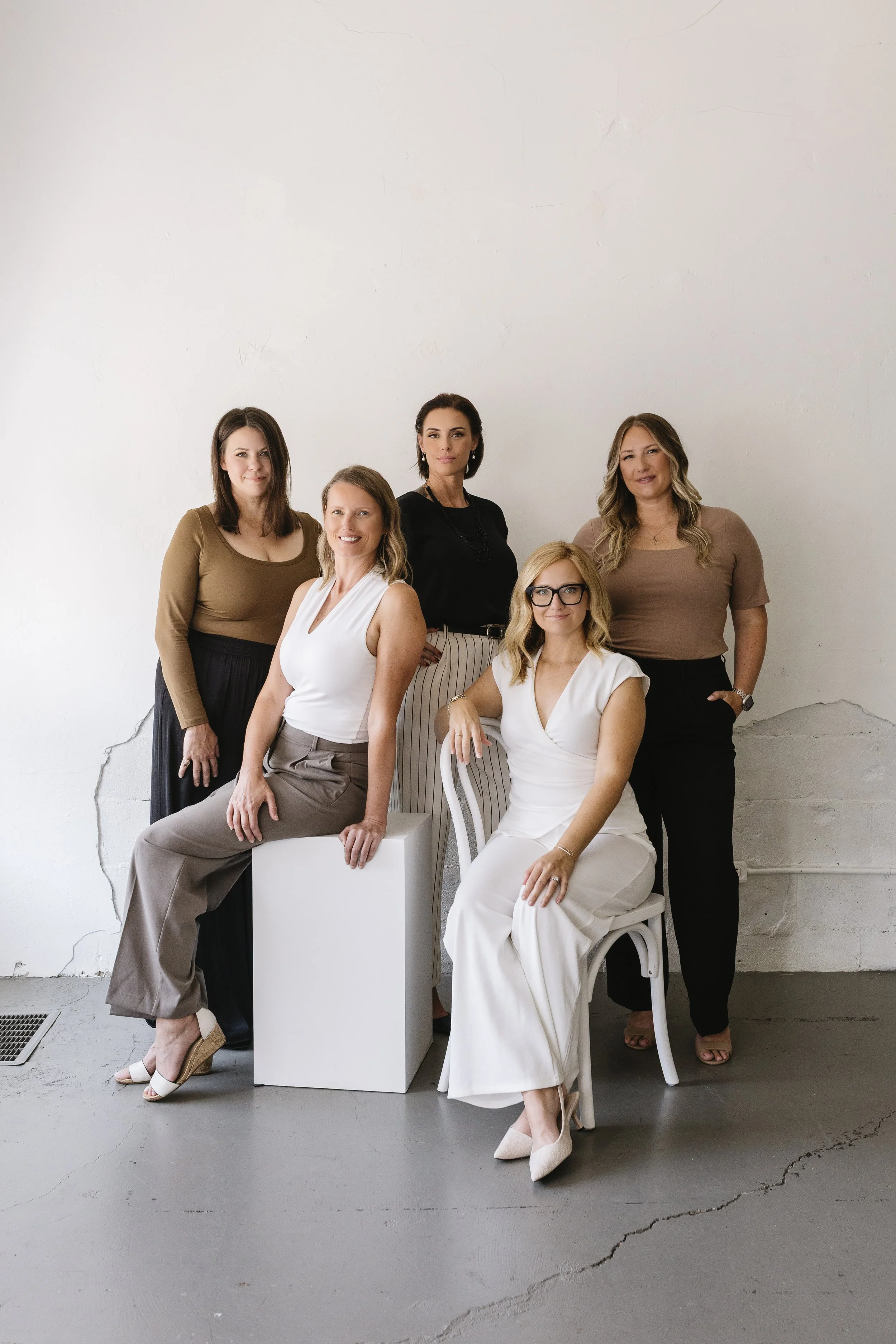 A group of five women from a bookkeeping firm posing against a plain white wall, with some sitting and some standing, dressed in professional attire.