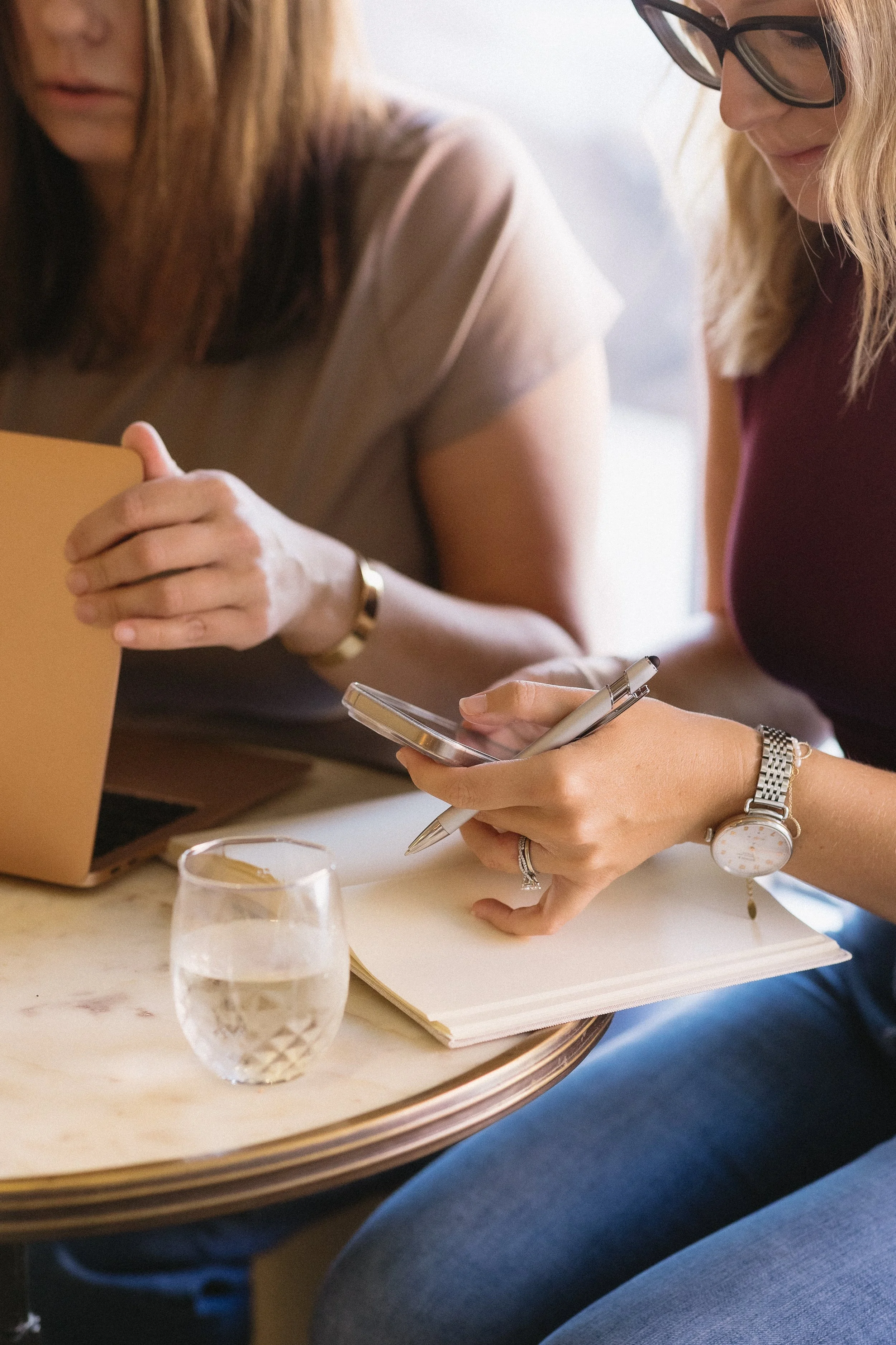 Two women sitting at a table, one is using a smartphone and writing in a notebook, with a glass of water nearby.