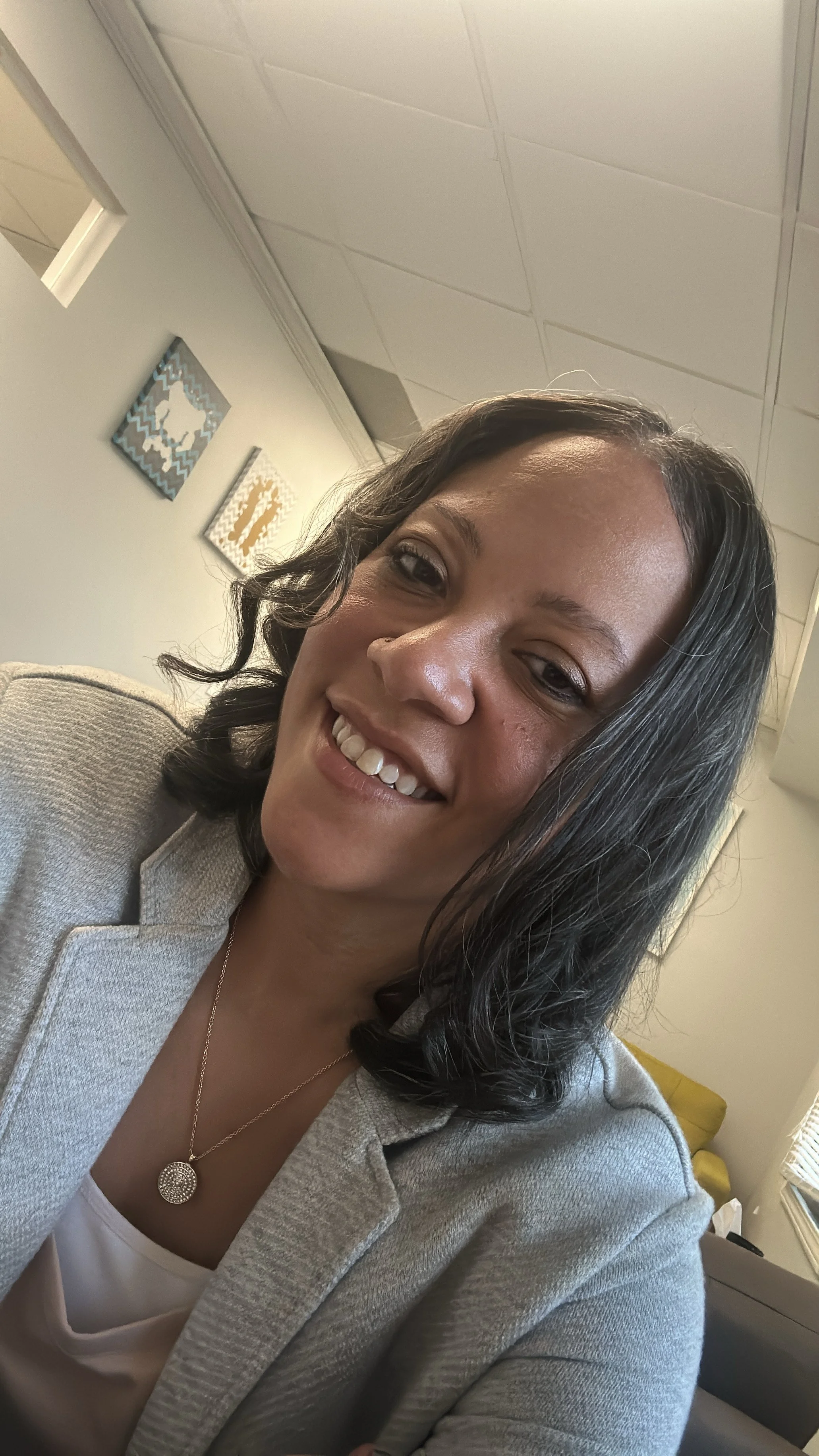 A woman with shoulder-length dark hair and a bright smile, wearing a light-colored blazer and a necklace, sitting in an indoor setting with wall art in the background.