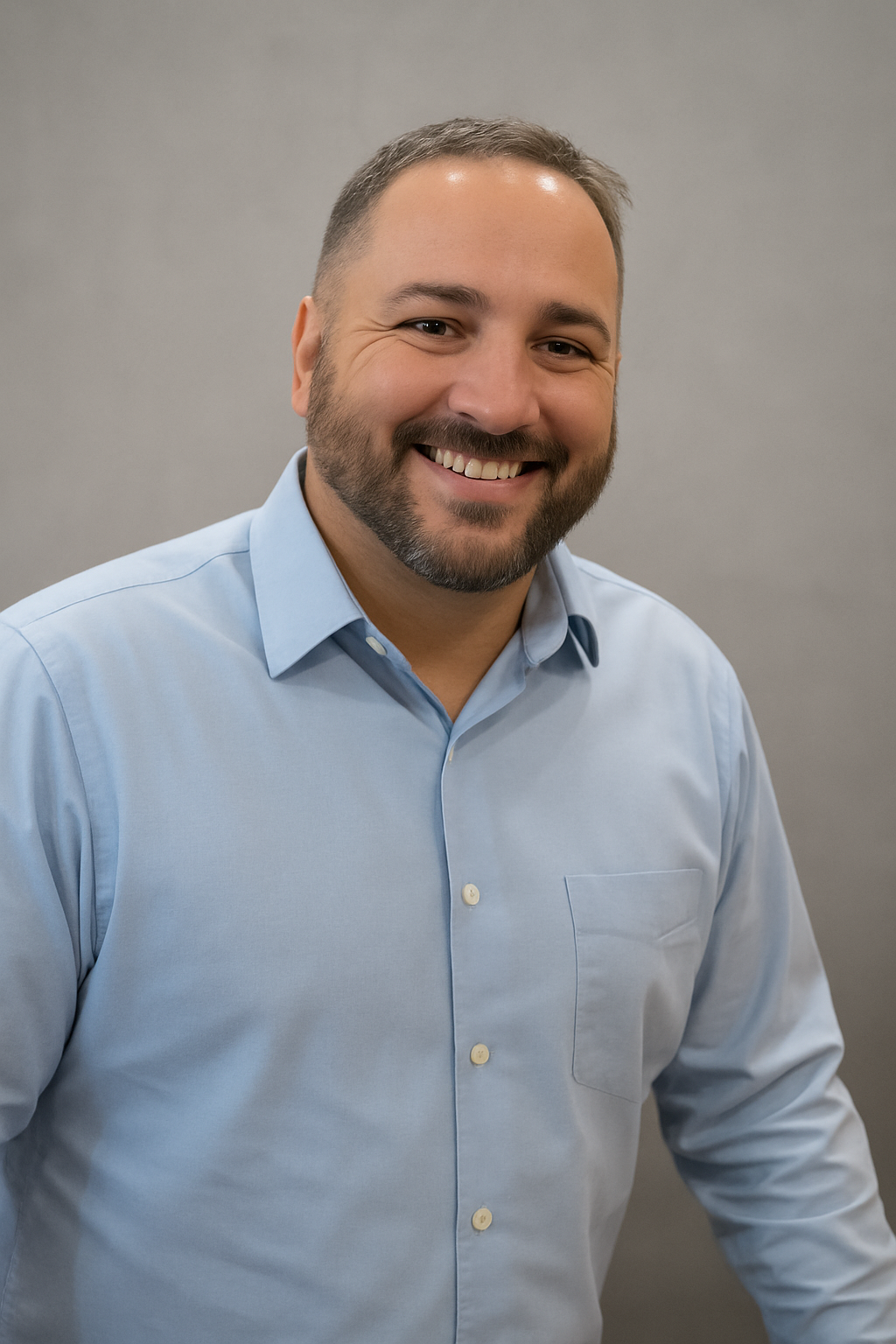 A smiling man with a beard and short hair, wearing a light blue dress shirt, standing against a plain gray background.