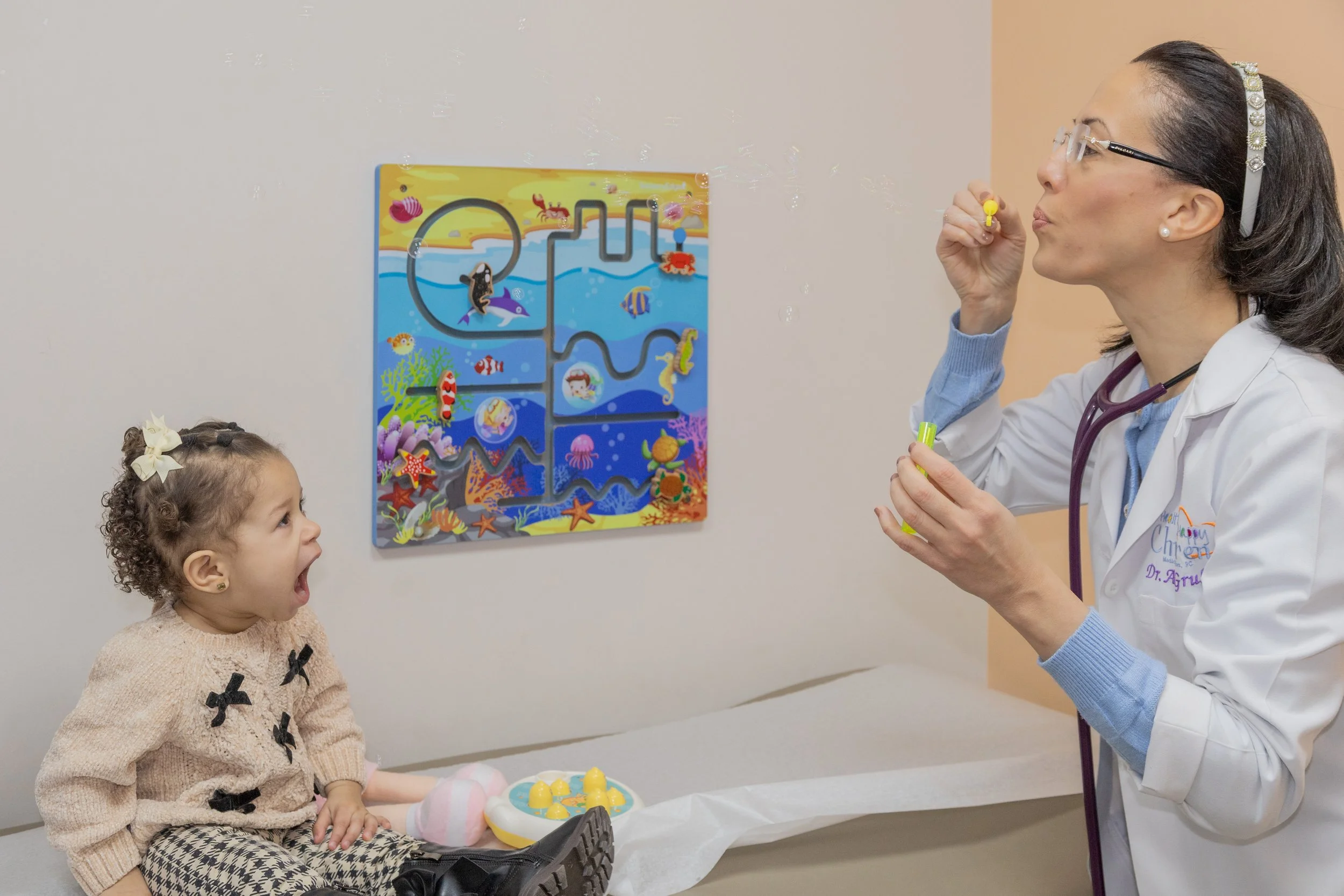 A young girl with curly hair and a bow on her head sits on an examination table, looking surprised as a female healthcare professional blows bubbles in a pediatric medical office.
