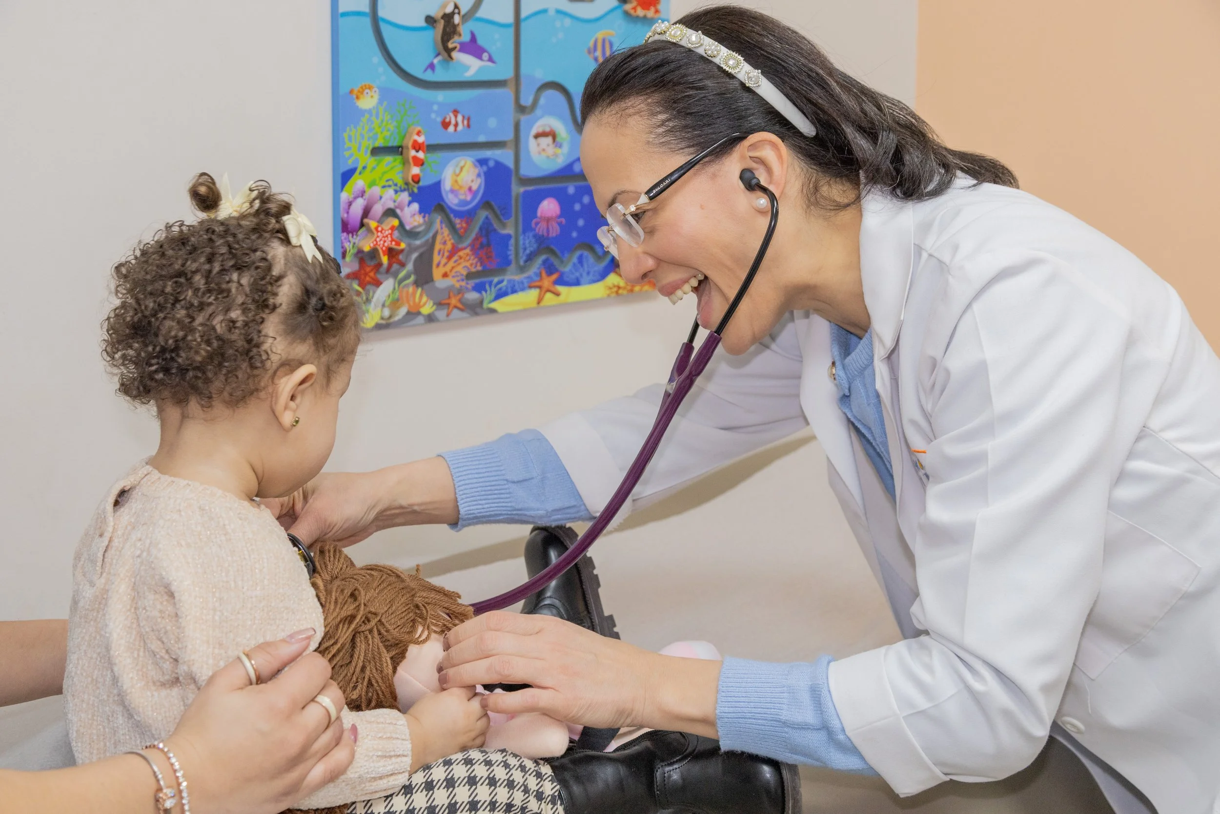 A female doctor using a stethoscope to listen to a young girl's chest during a check-up in a medical setting, with a colorful ocean-themed picture on the wall behind them.