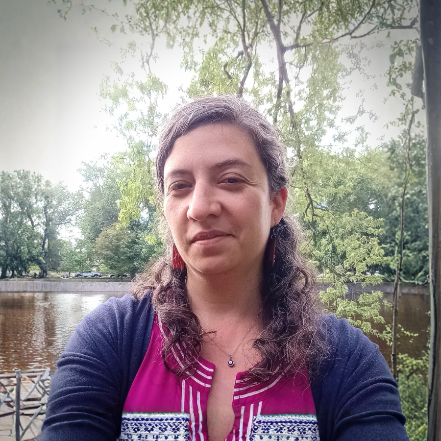 A woman with long wavy hair taking a selfie outdoors near a river with trees in the background.