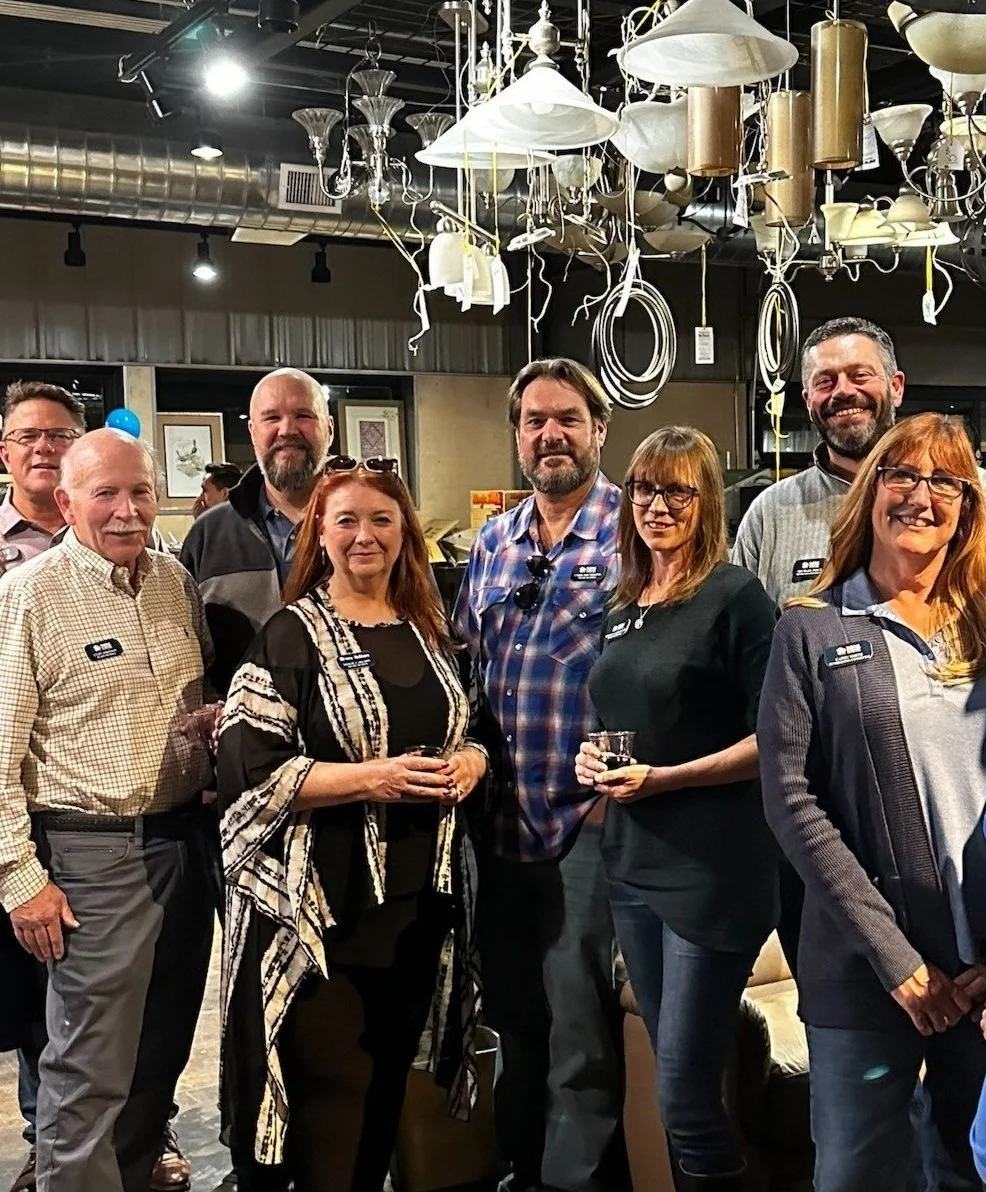 A group of eight people standing together indoors, smiling for a photo, with lighting fixtures hanging from the ceiling above them.