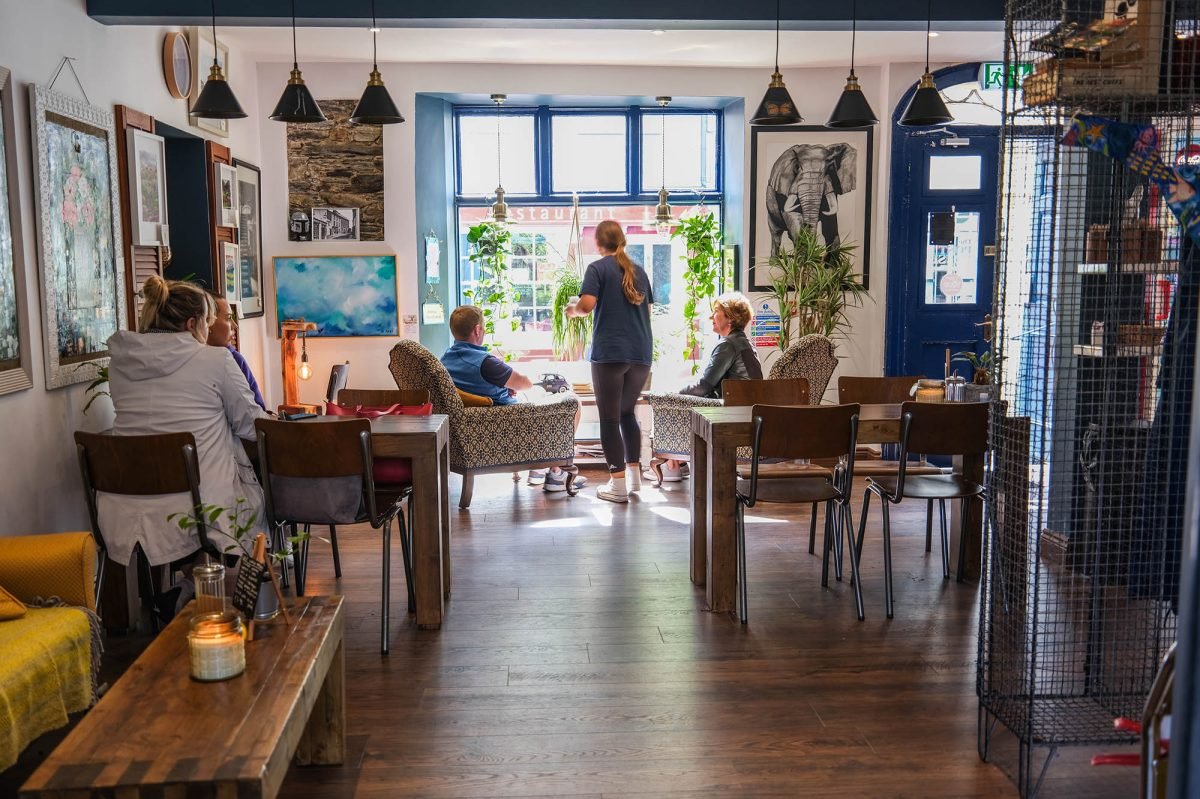 Interior of a cozy cafe with several patrons sitting and interacting, decorated with artwork and plants near a large window.
