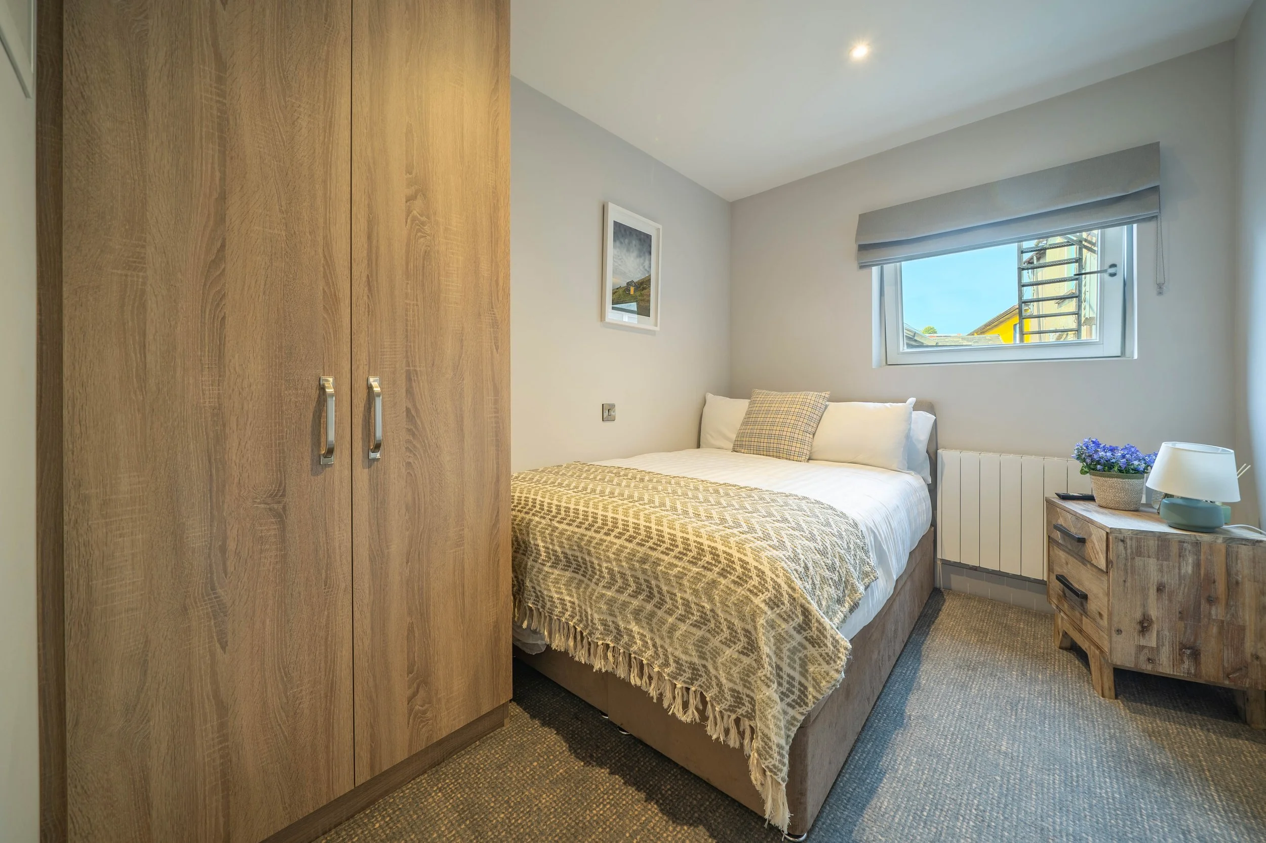 Cozy bedroom with a wooden wardrobe, bed with patterned throw blanket and pillows, beside a wooden nightstand with a flowerpot, lamp, and window showing a clear sky.