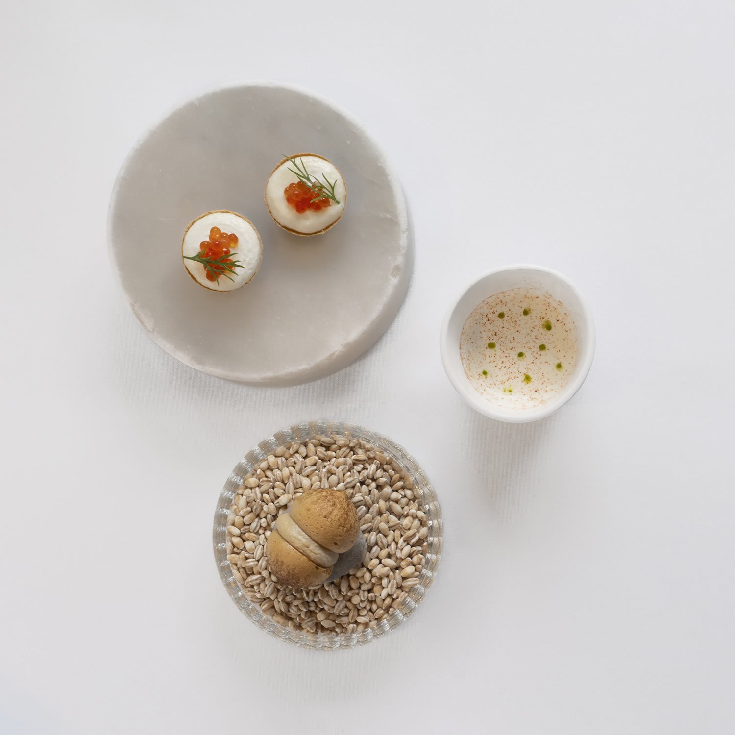 Two small savory pastries topped with red caviar and garnished with herbs on a white plate, a small bowl of frothy beverage with green droplets, and a glass dish with a round cookie and sunflower seeds.
