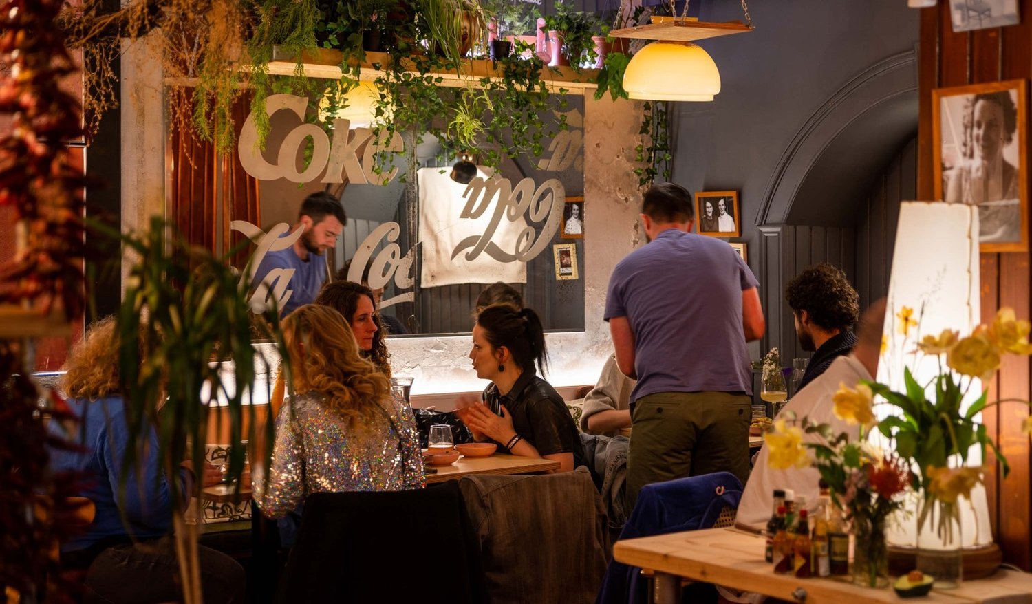People dining and socializing in a cozy restaurant with plants, framed photos, and warm lighting.