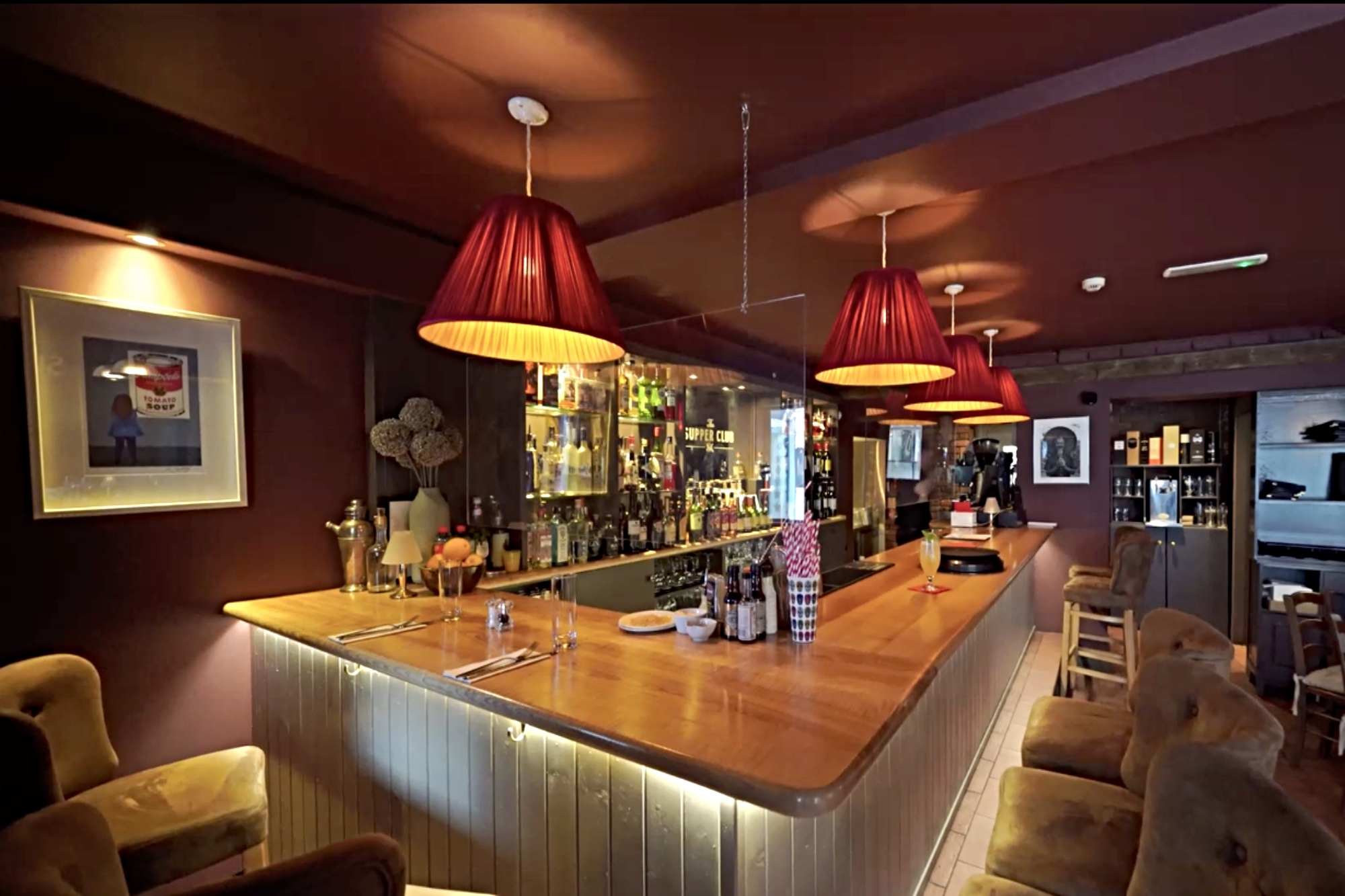 Interior of a cozy bar with a wooden counter, red pendant lamps, and plush chairs, with shelves of bottles and framed pictures on the walls.