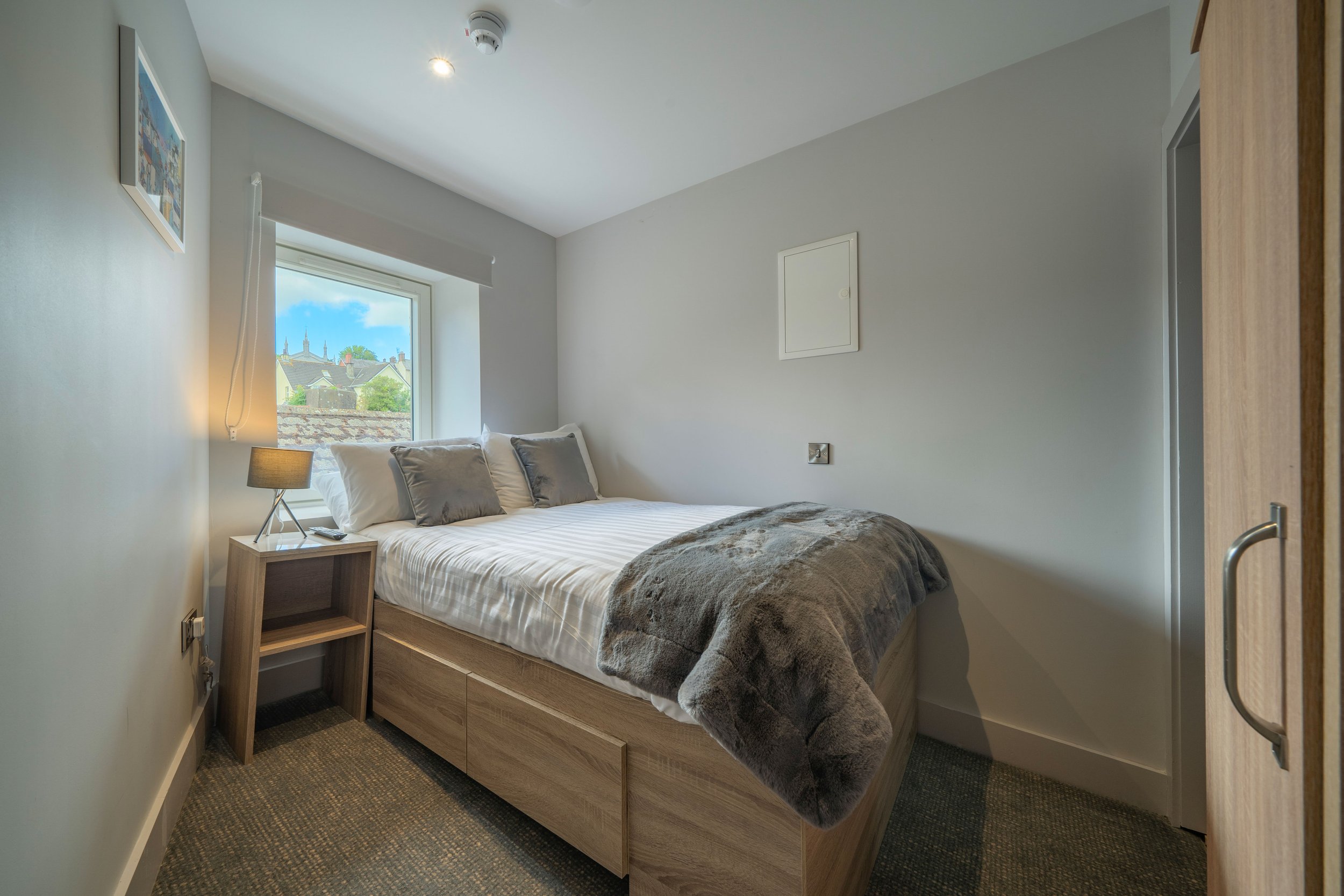 Clean bedroom with a window, bed with white and gray bedding, wooden bedside table with lamp, and a gray fur throw blanket.