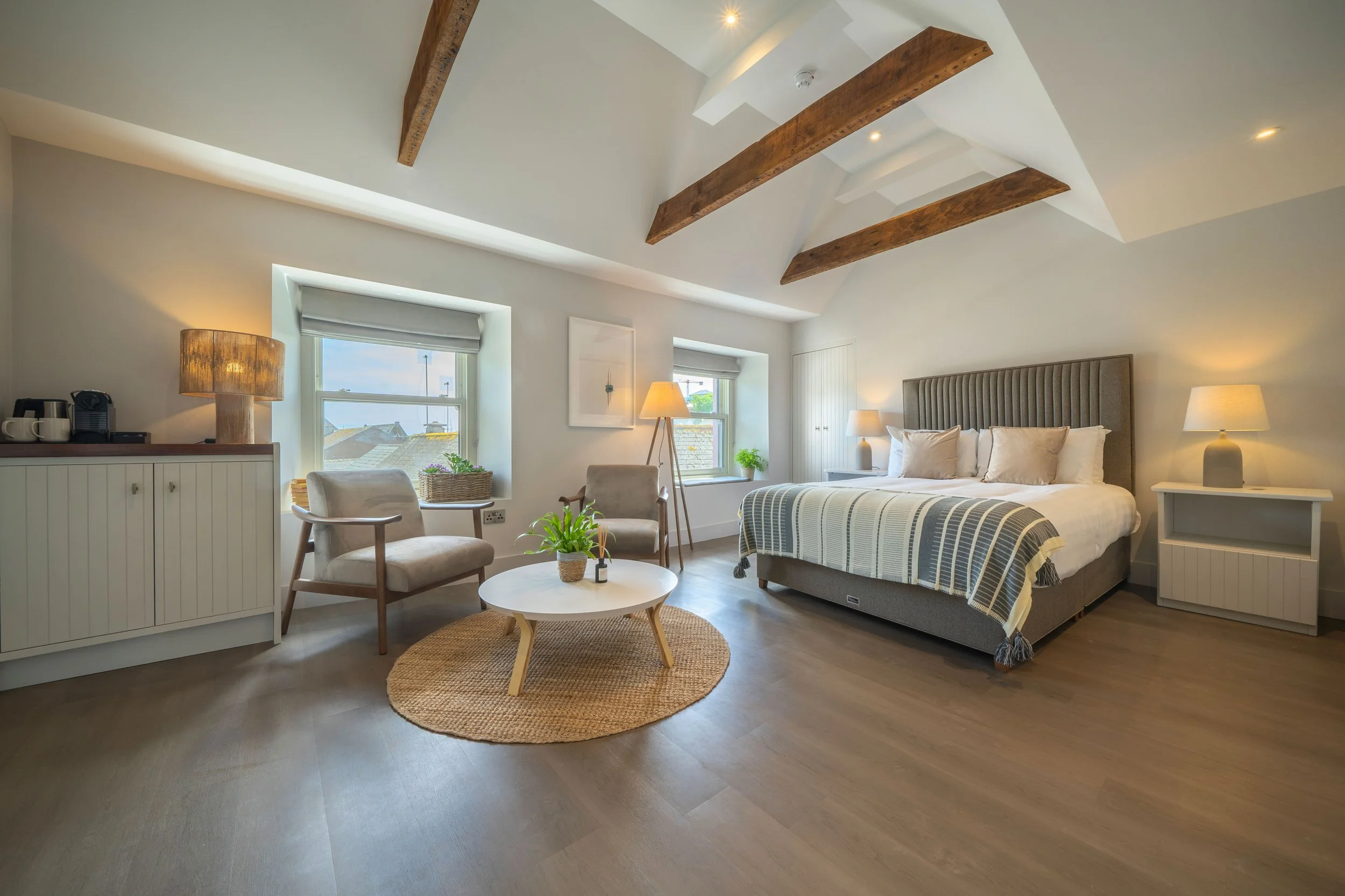 Bright and modern hotel bedroom with a large bed, sitting area with two chairs and a round coffee table, two windows with views, and decorative wooden beams on the ceiling.