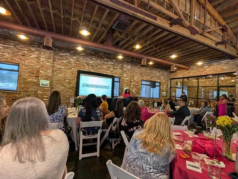 People sit around tables at a morning breakfast event for a local Tacoma nonprofit. There is a speaker wearing a pink dress.