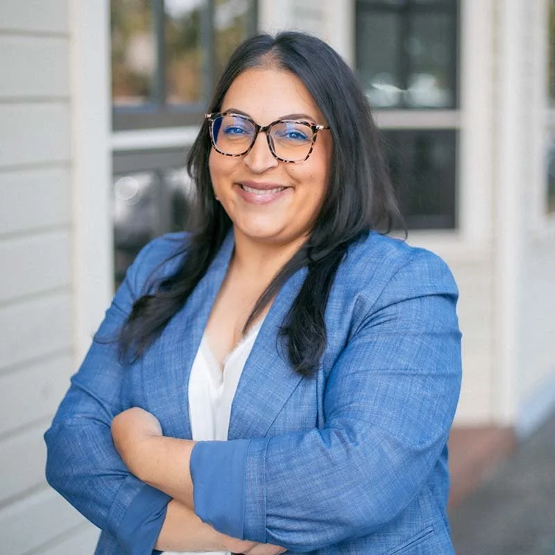 Woman in a blue blazer jacket smiles at the camera. She has glasses.