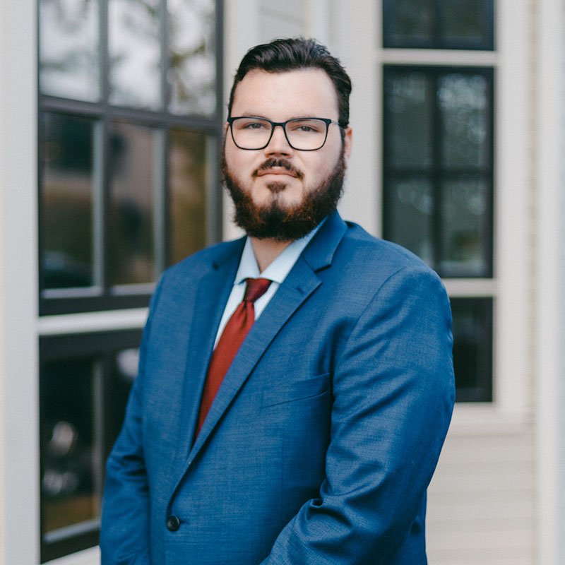 Bearded man in glasses with dark hair, slightly smiles at camera wearing a blue suit with red tie