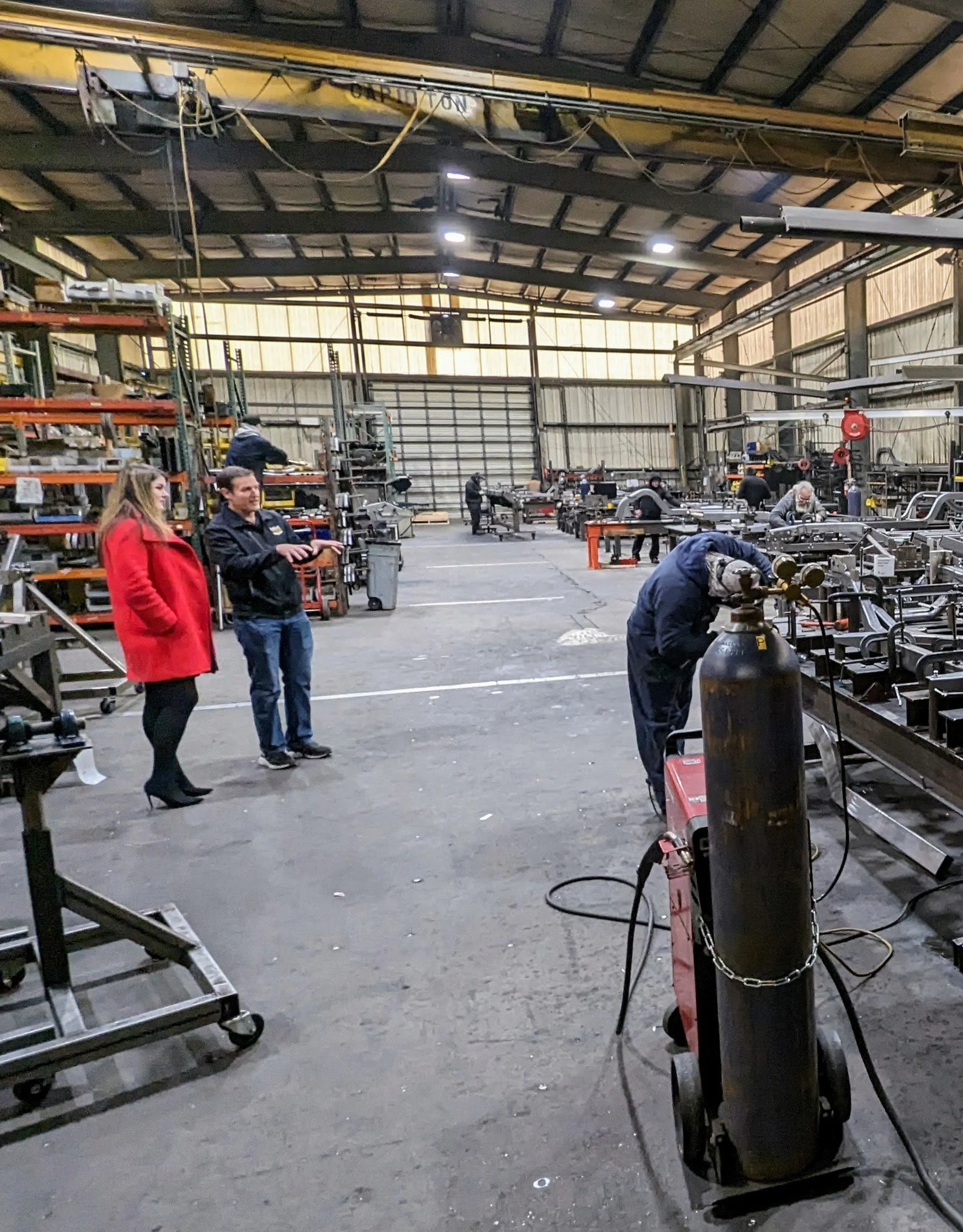 Brooke Johnson, in a red coat, talks with Matt Jones of Art Morrison Enterprises in their fabrication shop. Welding tools and supplies are visible.