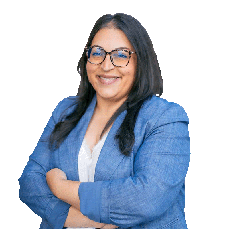 A woman with long dark hair, glasses, and a blue blazer smiling with arms crossed against a white background.