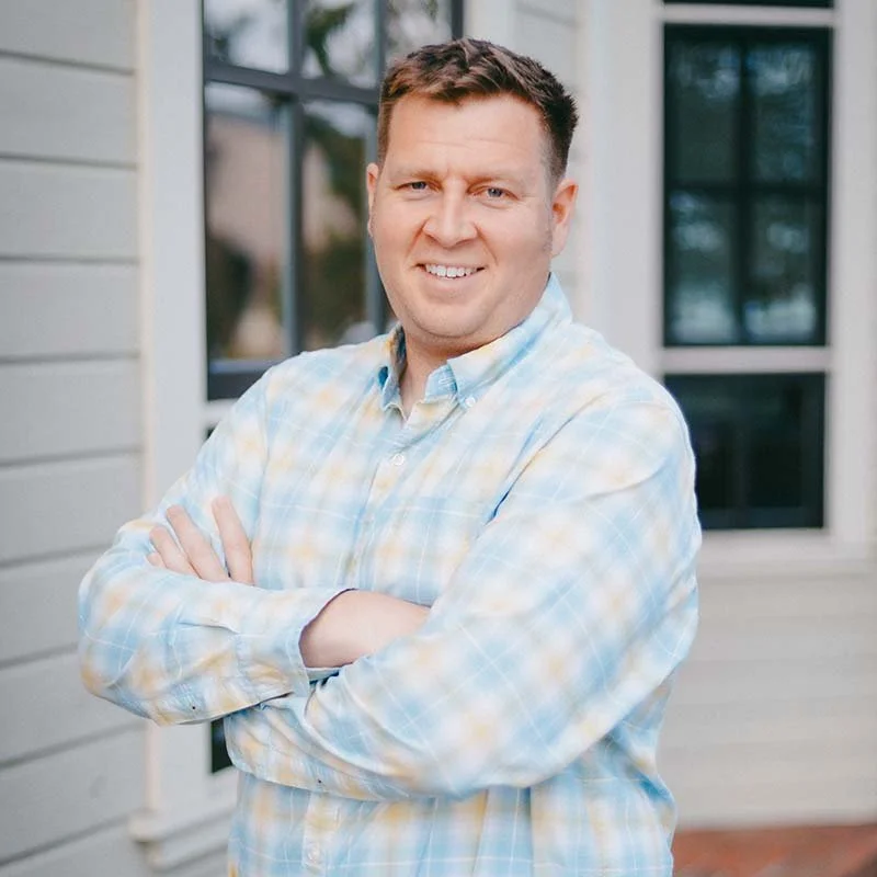 Man smiles at camera with his arms crossed. He's in a light blue checkered dress shirt.