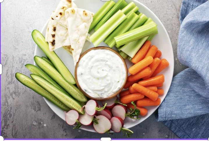 A white plate filled with sliced cucumbers, radishes, baby carrots, celery sticks, pita bread triangles, and a bowl of ranch dressing in the center.