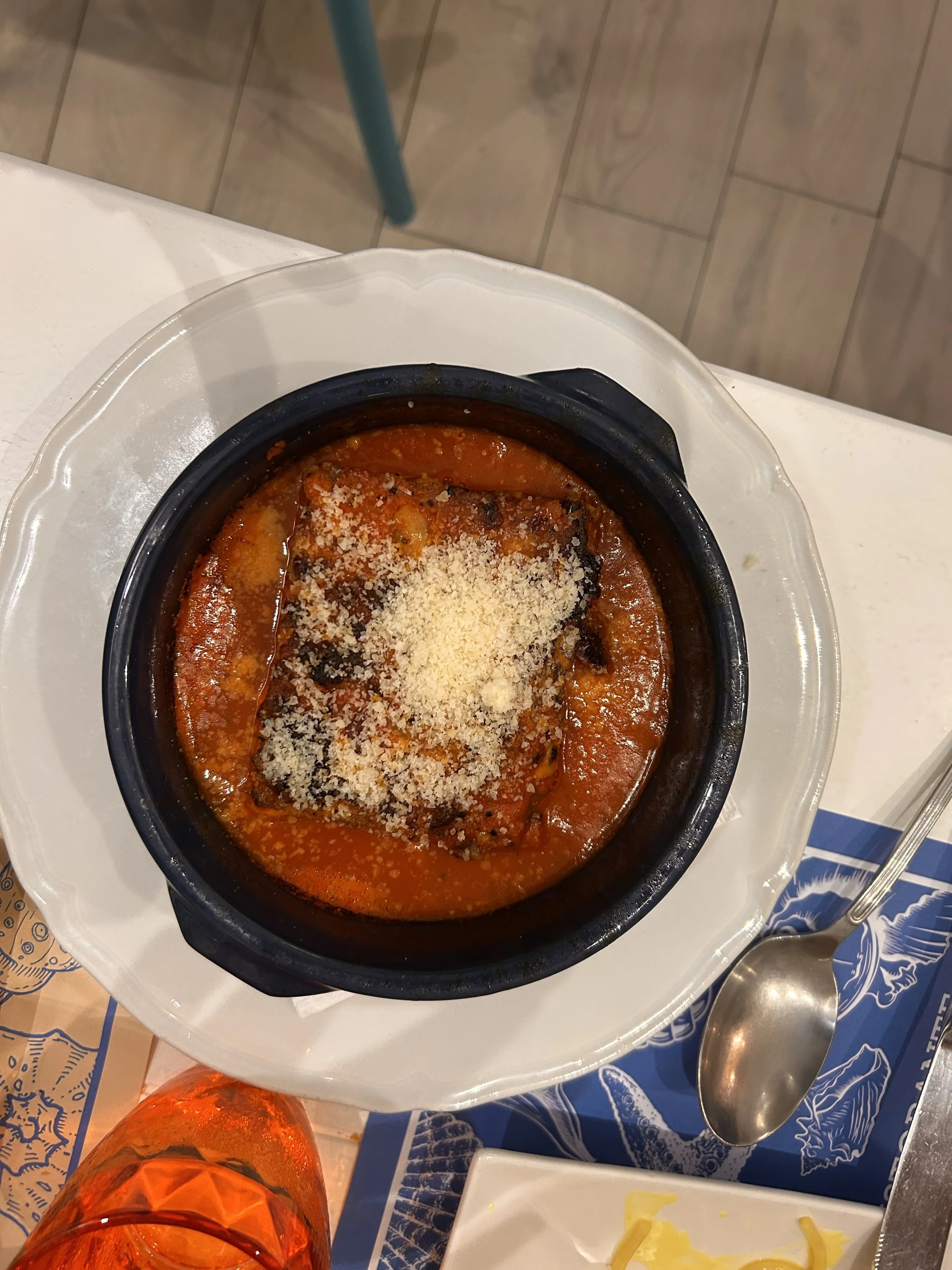 Baked eggplant parmesan in a black bowl topped with grated cheese, on a white plate with a spoon nearby.