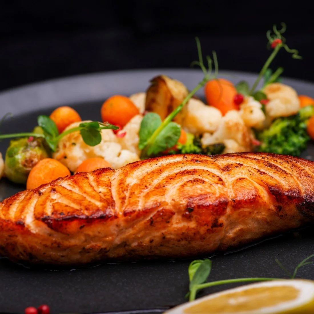 Grilled salmon with vegetable medley including cauliflower, carrots, and broccoli, garnished with greens, on a black plate.