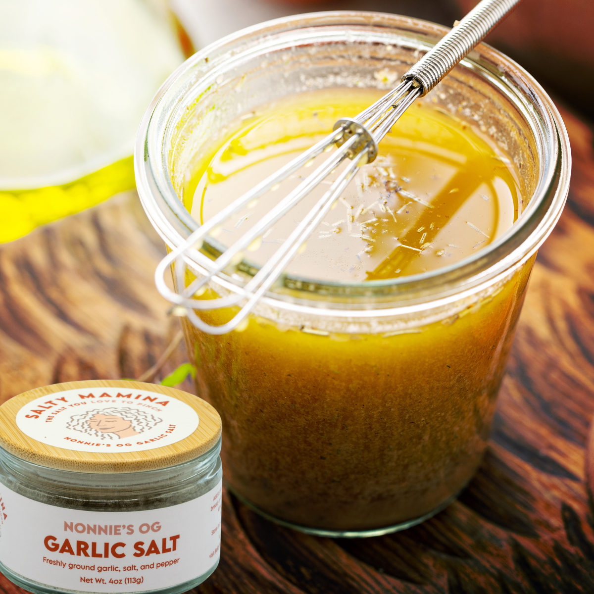 A glass jar of homemade salad dressing made with SALTY MAMINA garlic salt with a metal whisk resting inside, placed on a wooden surface. Next to the jar is a container labeled "Nonnie's OG Garlic Salt" with a red and white label.