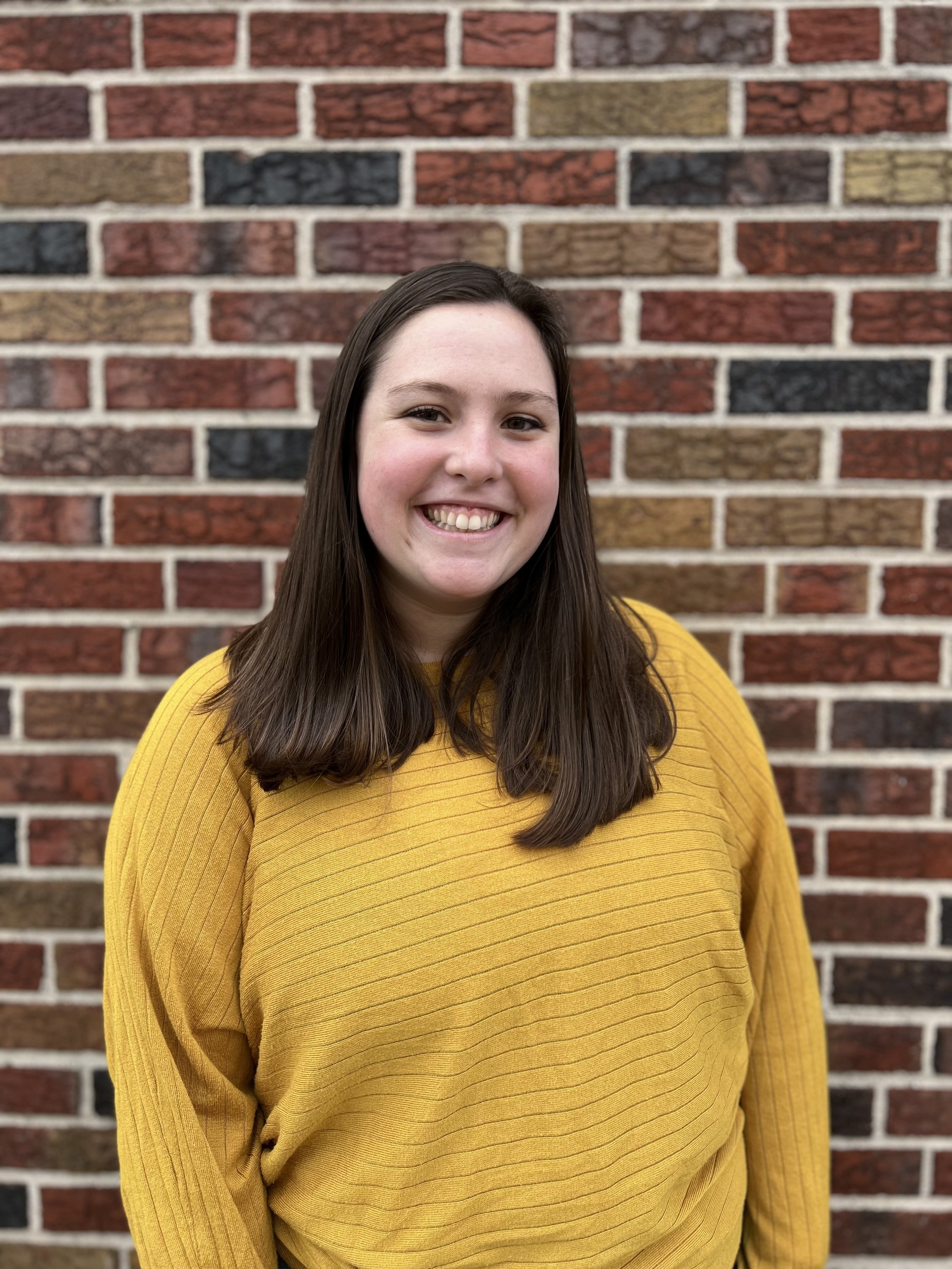 Person in a yellow sweater smiling in front of a multicolored brick wall.
