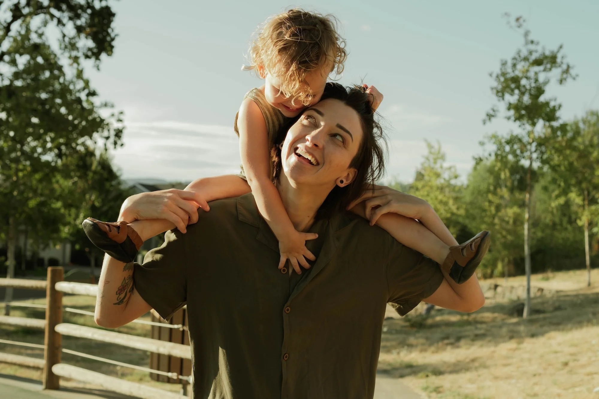 Mother carrying child on shoulders outdoors, smiling in warm golden light