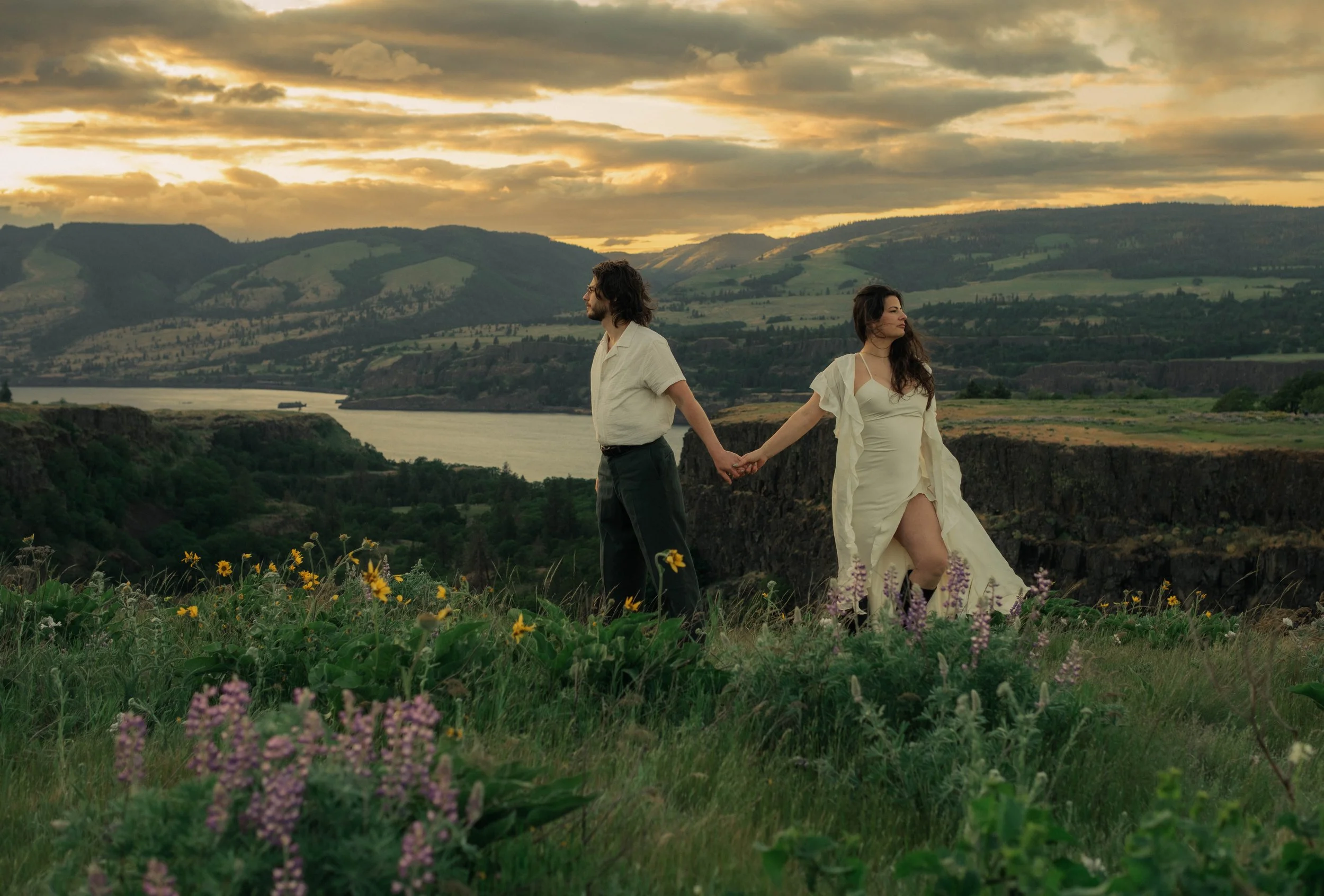 Couple holding hands in wildflower field overlooking river and mountains at sunset